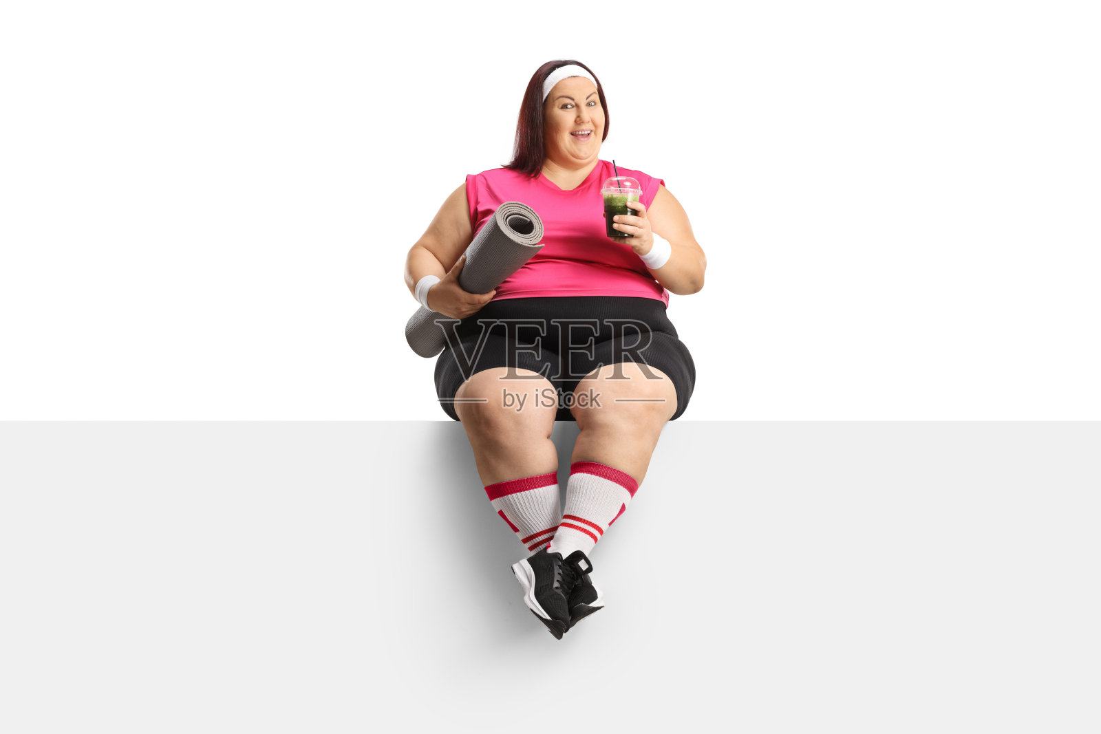 穿着运动服的大码女人坐在一块空白的面板上，手里拿着一个运动垫和一杯奶昔照片摄影图片