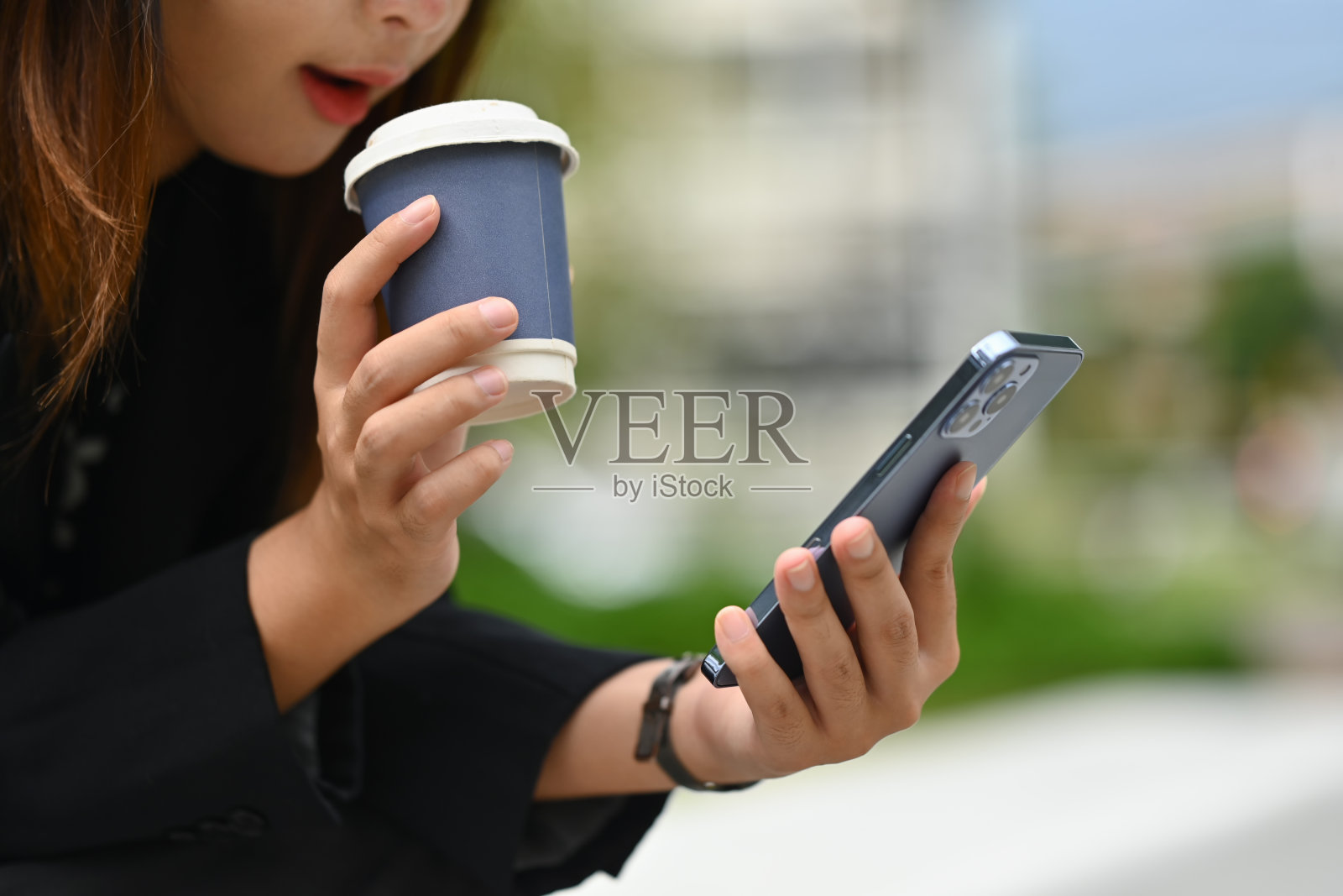 阳光明媚的一天，拿着外卖咖啡的年轻女子在办公大楼外用手机照片摄影图片