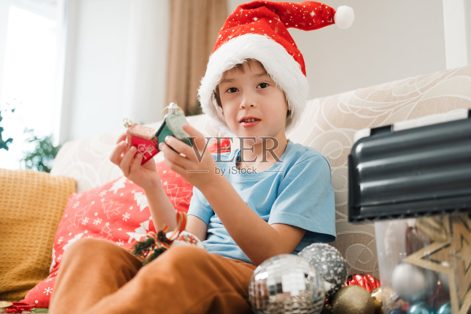 戴着红色圣诞帽的快乐微笑的男孩正坐在客厅的沙发上玩圣诞球，准备装饰圣诞树。照片摄影图片