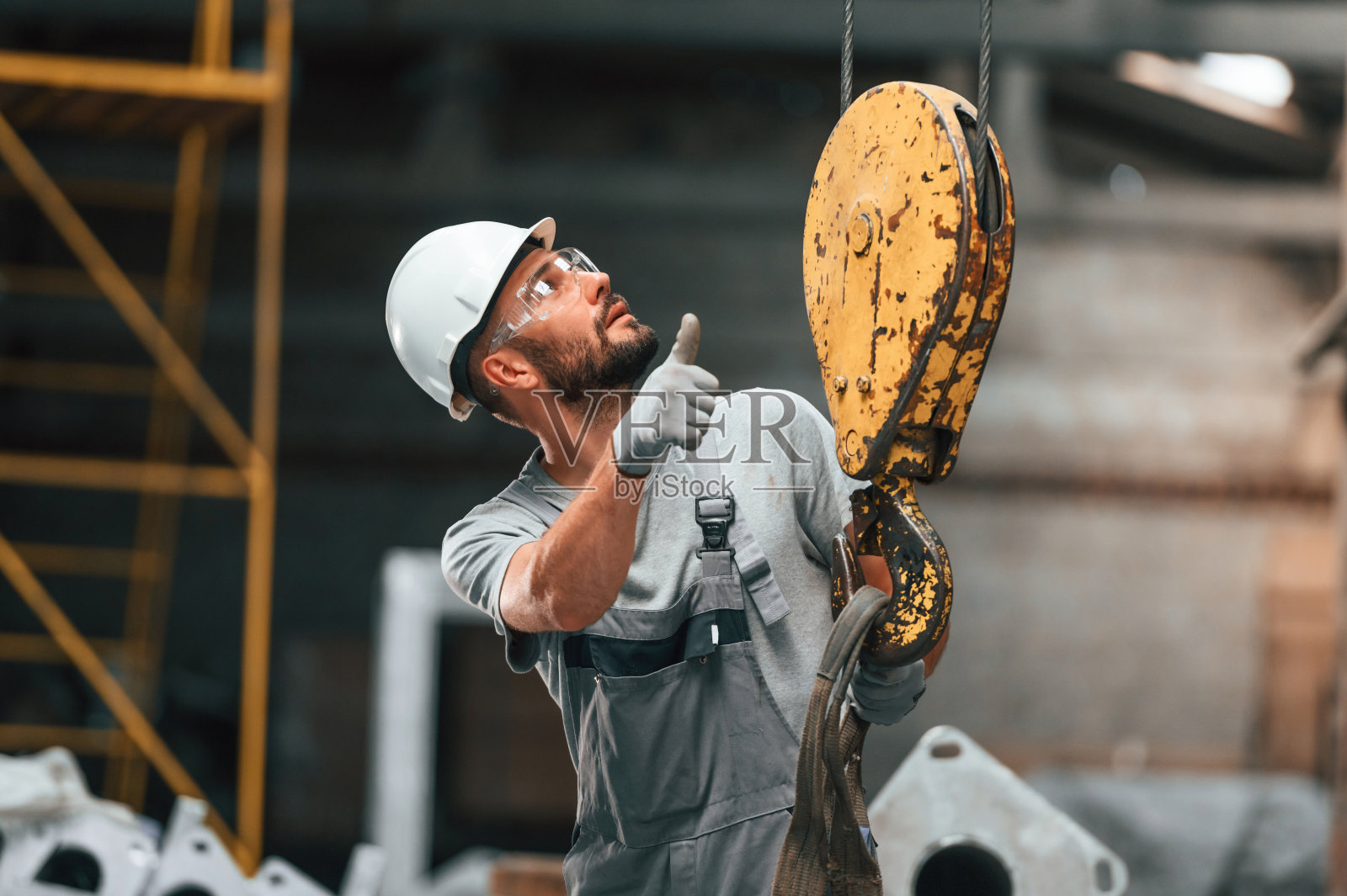 黄色吊车钩。穿着灰色制服的年轻工厂工人照片摄影图片