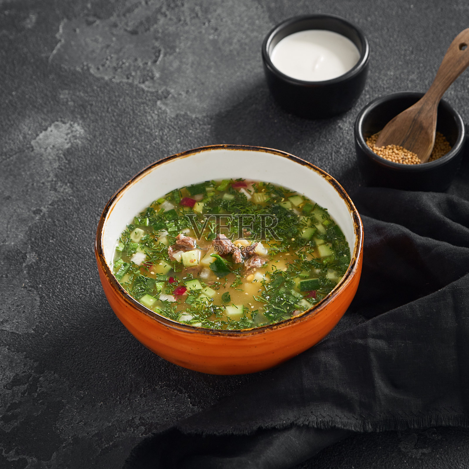 传统俄式冷汤配牛肉，盛在橙色碗中，深色石板背景，附近有酸奶油和香料照片摄影图片