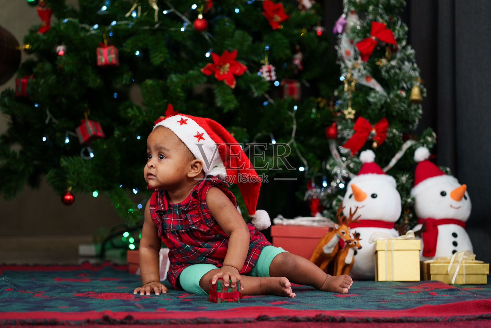 快乐的非裔美国女婴穿着圣诞老人的服装，微笑着坐在圣诞树前，拿着礼物盒照片摄影图片