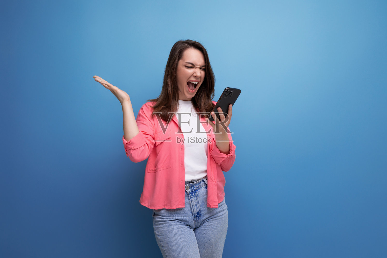 惊讶的年轻女子穿着粉色衬衫和牛仔裤，手机在工作室孤立的背景照片摄影图片