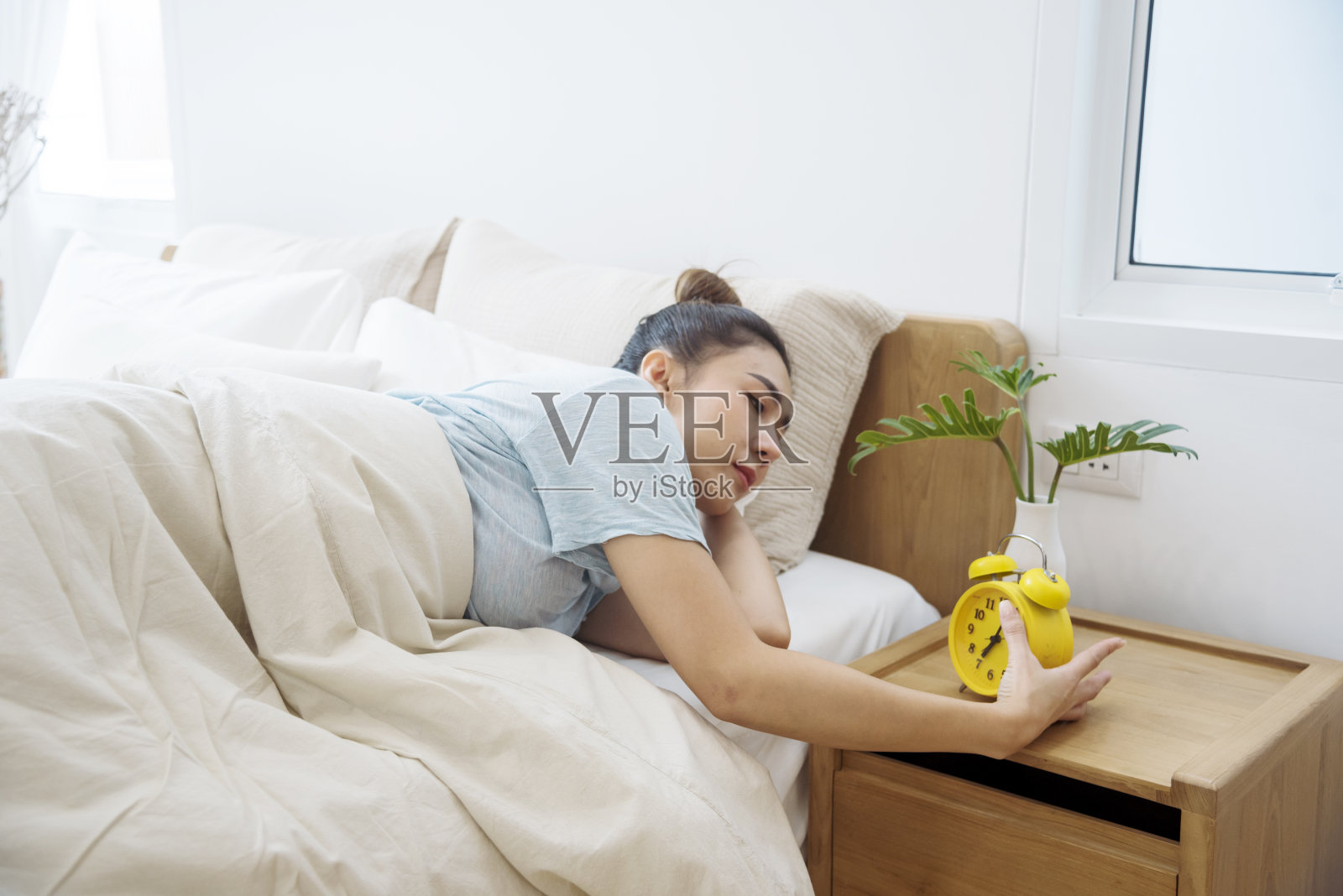 亚洲女性在白色舒适的卧室里醒来，享受清晨日出的快乐时光。美丽的中国人使用手动停止时间闹钟。快乐的人生活方式早上醒来健康放松困倦打盹照片摄影图片