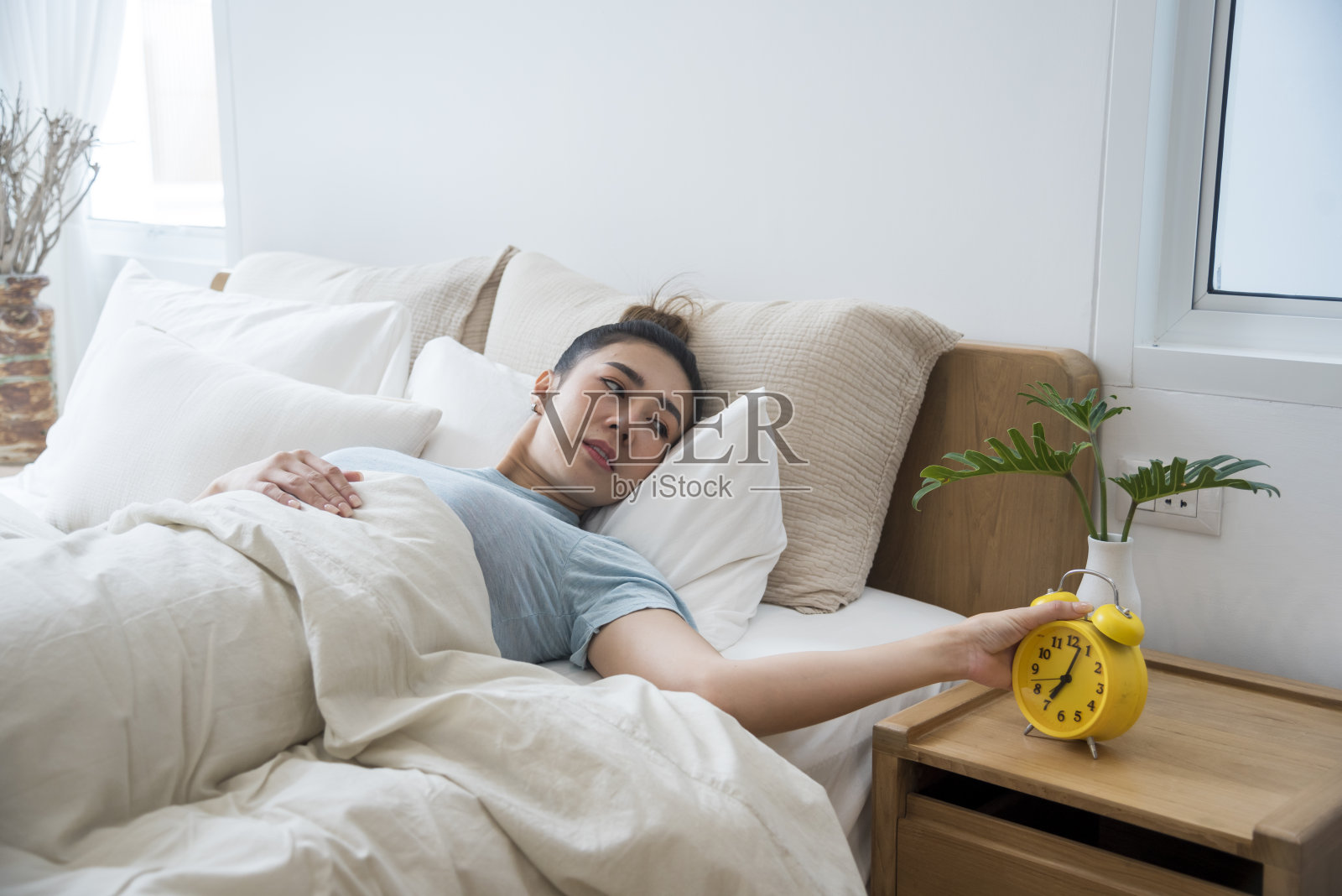 亚洲女性在白色舒适的卧室里醒来，享受清晨日出的快乐时光。美丽的中国人使用手动停止时间闹钟。快乐的人生活方式早上醒来健康放松困倦打盹照片摄影图片