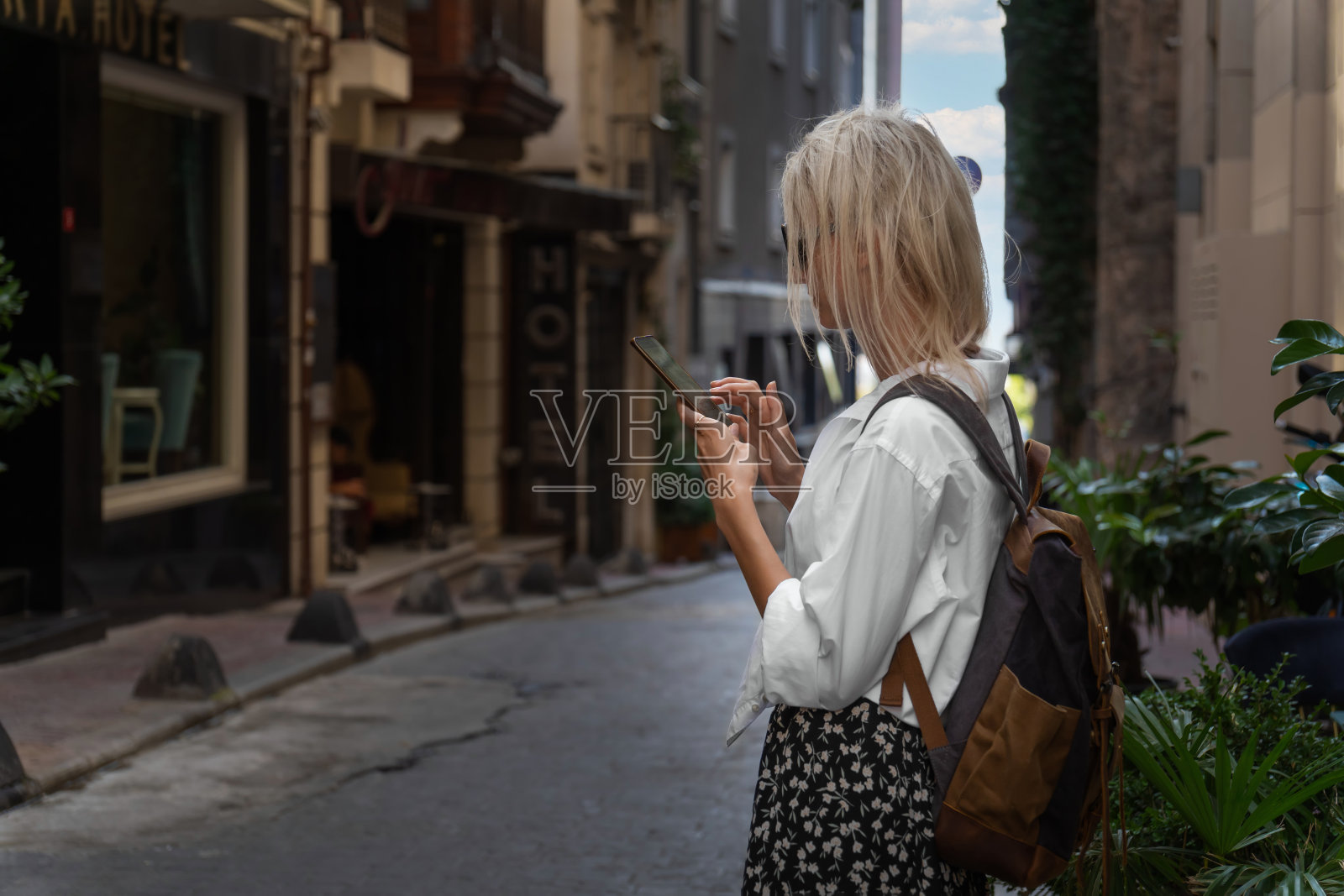 年轻的金发女游客穿着白衬衫和裙子，背着手工背包，手里拿着手机站在伊斯坦布尔一条美丽的街道上。旅游理念，现代科技。照片摄影图片