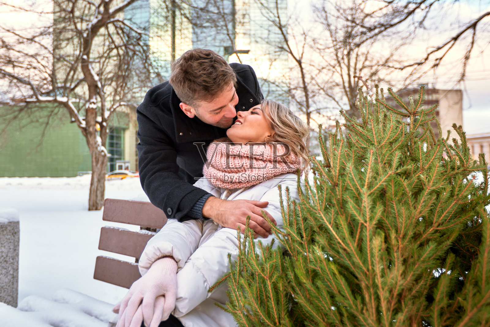 一对美丽的年轻情侣坐在圣诞市场的长椅上，刚刚买了一棵圣诞树。男人和女人正在为圣诞节做准备。在大雪纷飞的城市里，人们期待着新年的到来。照片摄影图片