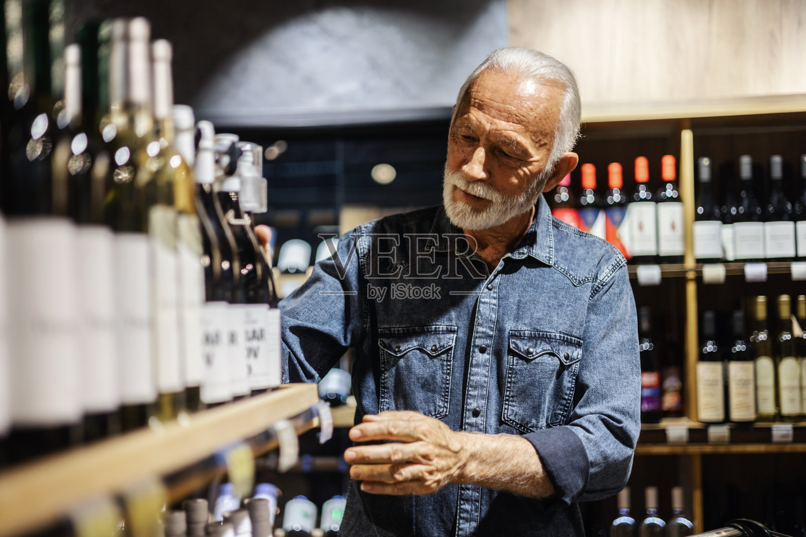 男性消费者在酒庄商店购买一瓶红酒。微笑着的成熟男子穿着休闲的衣服在超市杂货店购物，在大卖场里买酒选酒。照片摄影图片