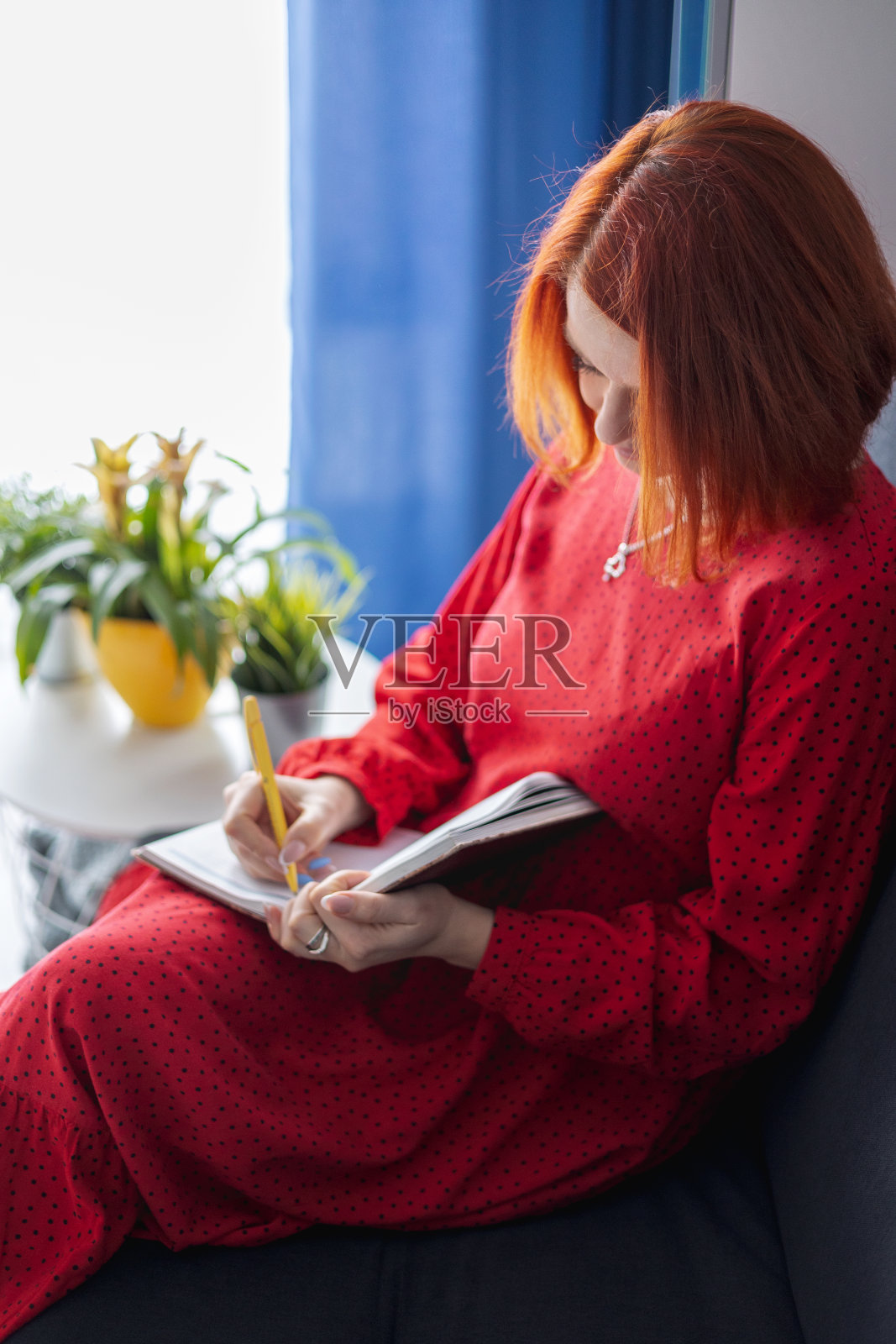 红头发的年轻女子坐在沙发上，在笔记本上做笔记。照片摄影图片