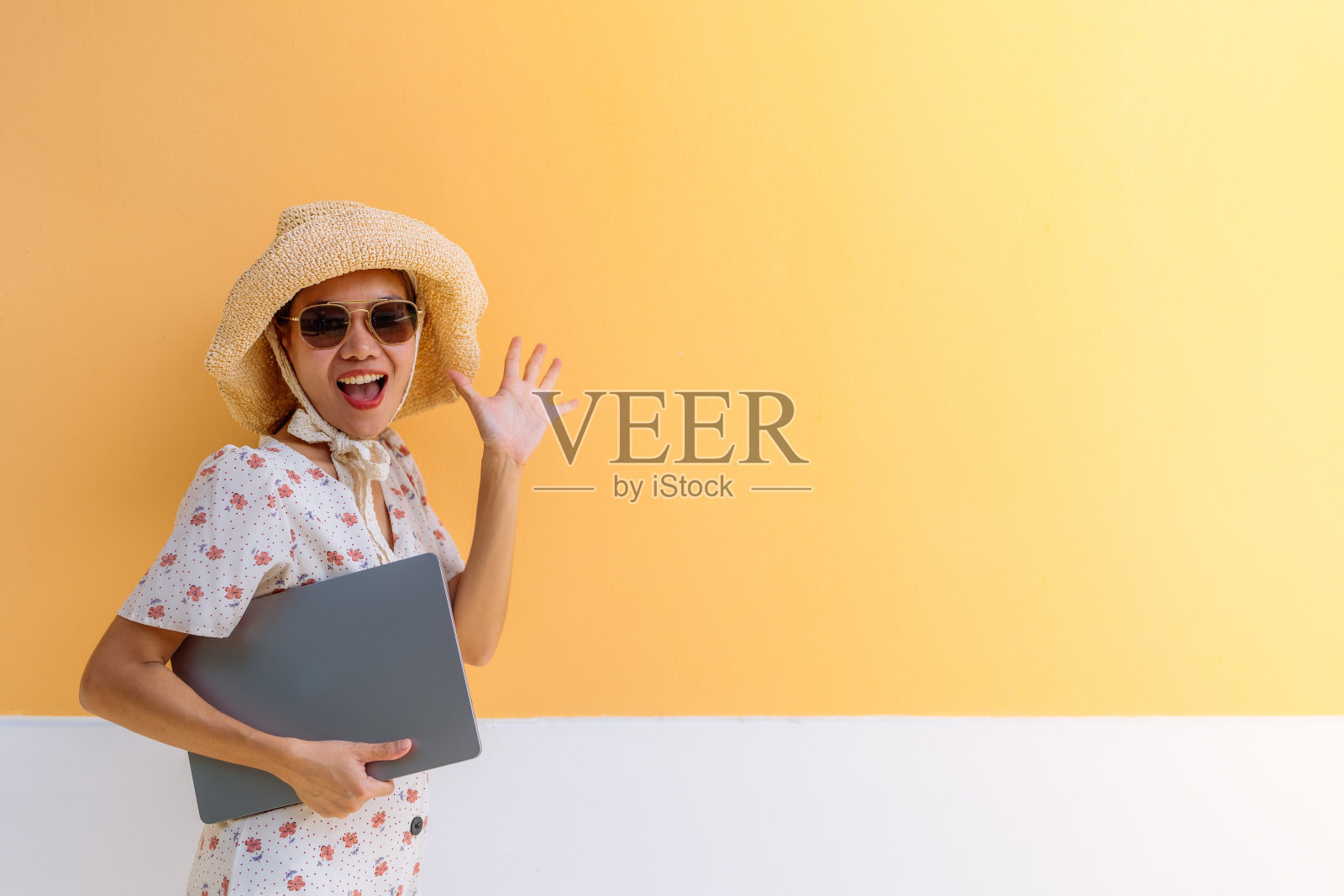 亚洲女性商务游客身穿花裙，戴着帽子，戴着眼镜，拿着笔记本电脑，她很高兴地举起了手，象征着数字5。背景是一面橙色的墙。照片摄影图片