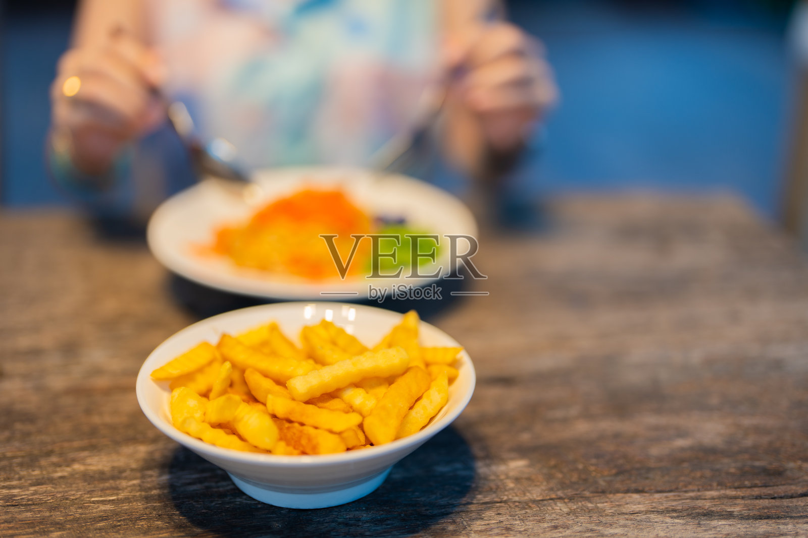 一大碗炸薯条菜单的特写照片，由著名厨师在餐馆的桌子上制作。背景里，一个女人正在津津有味地吃着面条。在温暖的灯光下照片摄影图片