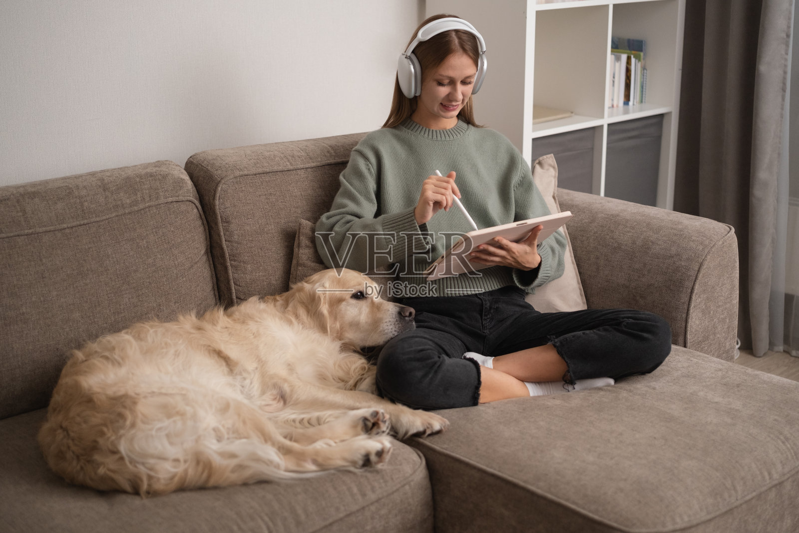 女孩戴着耳机和她的狗坐在沙发上。一位年轻女子在家工作，旁边是她的狗。照片摄影图片