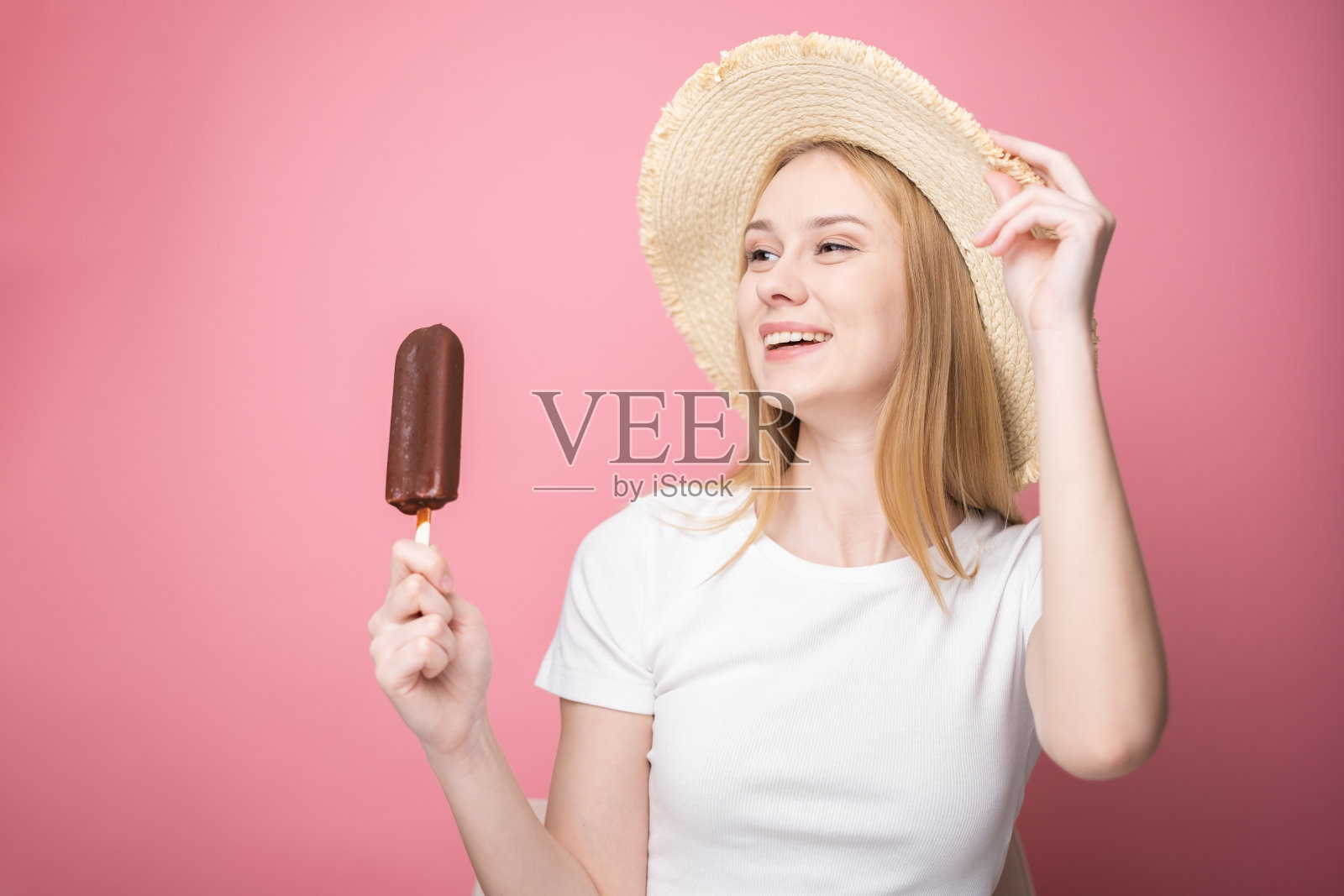 快乐的年轻女子戴着草帽，站在粉红色的背景上吃冰棒。夏日、度假、热天冷餐的概念。照片摄影图片
