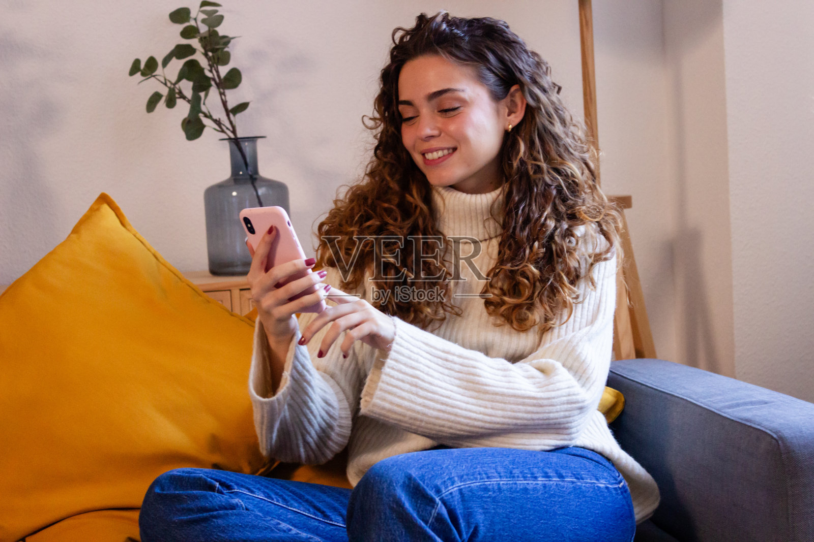 微笑的年轻女子坐在家里的沙发上玩手机照片摄影图片