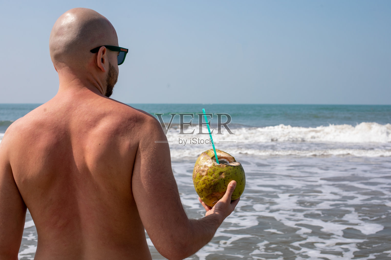 一名戴墨镜的男子站在海边的海浪中，背对着镜头，右手拿着剥了皮的椰子。照片摄影图片
