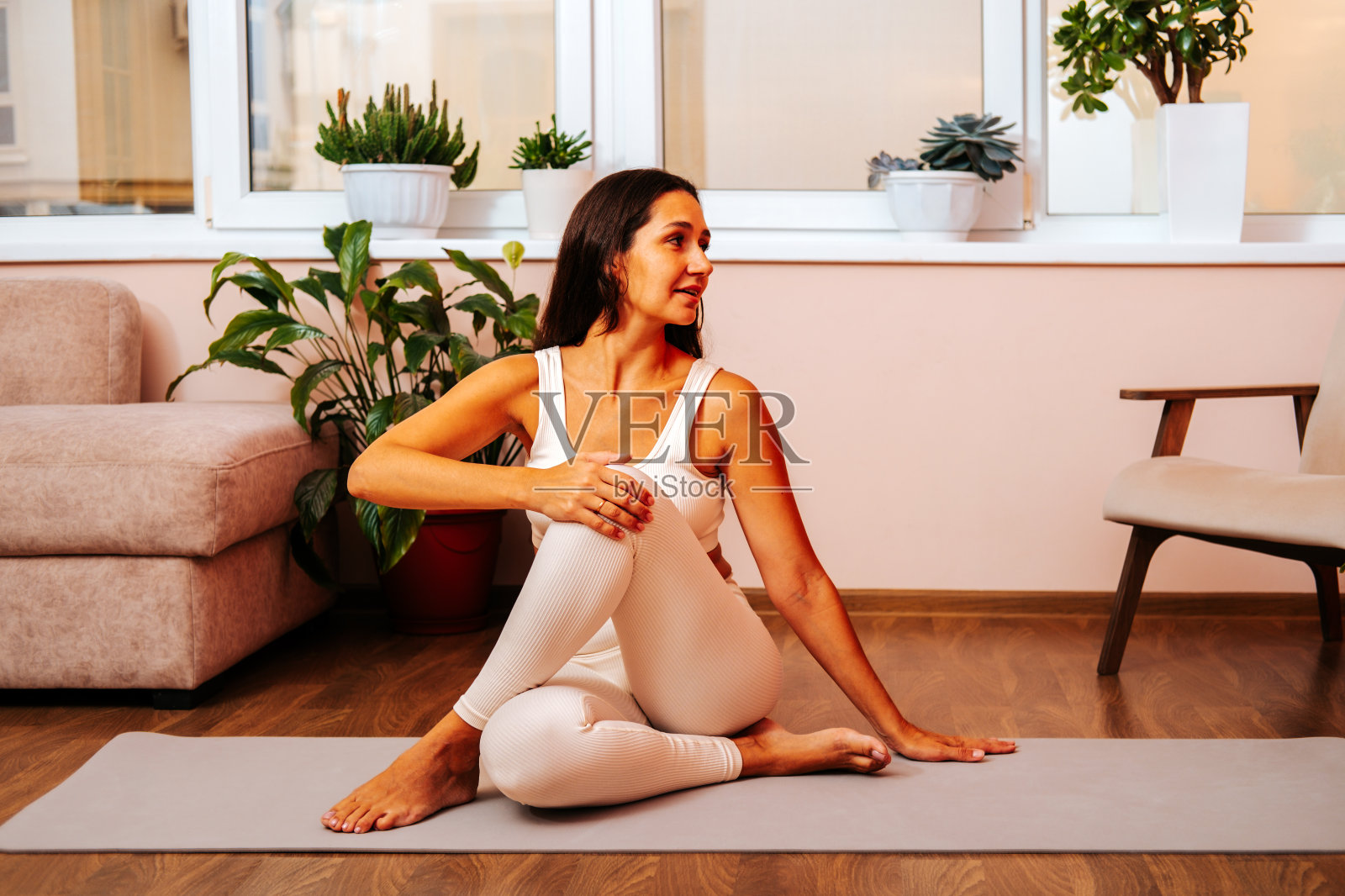 漂亮的年轻女子穿着白色运动服坐在瑜伽垫上，在明亮的现代客厅。瑜伽与健康习惯的概念。照片摄影图片