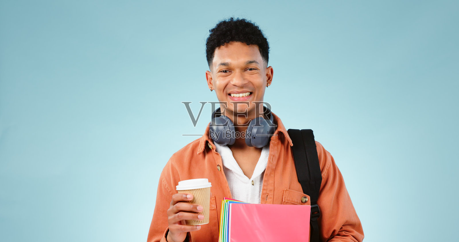 在大学或学院的工作室里，学生、咖啡和拿着书和背包的男人的肖像。教育，快乐和人的微笑，耳机，笔记本和蓝色背景的学习袋照片摄影图片
