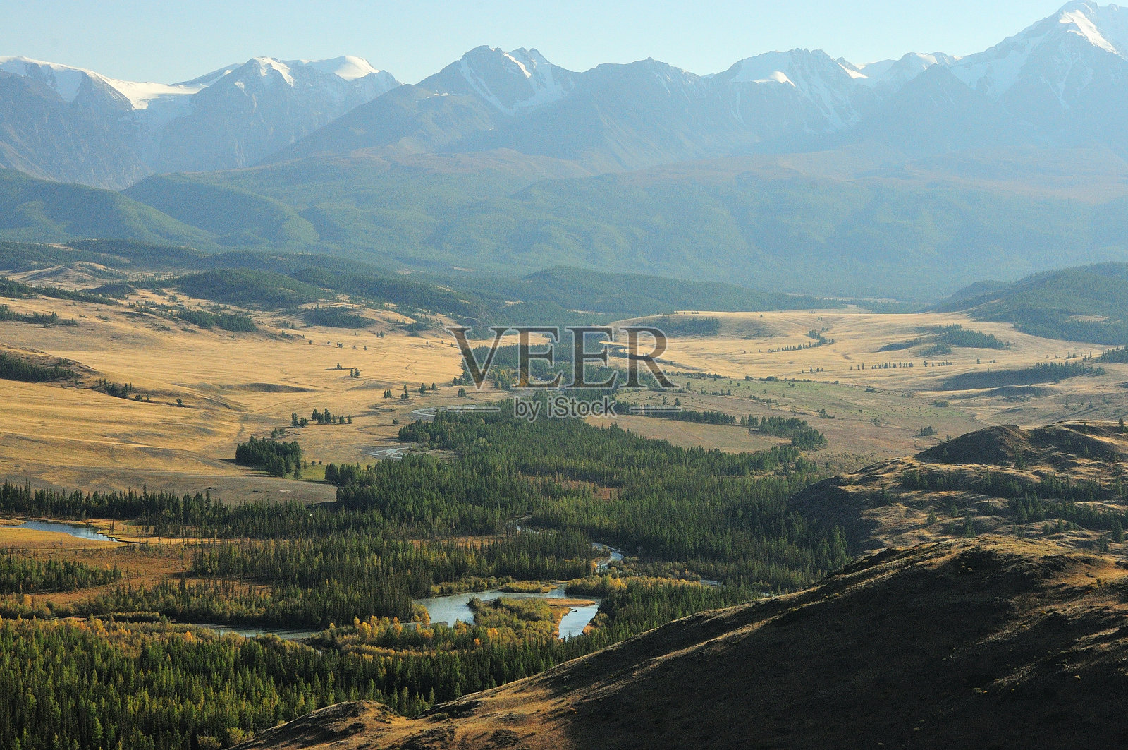 在落日的余晖中，一条美丽的河流蜿蜒曲折，两岸树木繁茂，流经山脚下的秋季草原。照片摄影图片