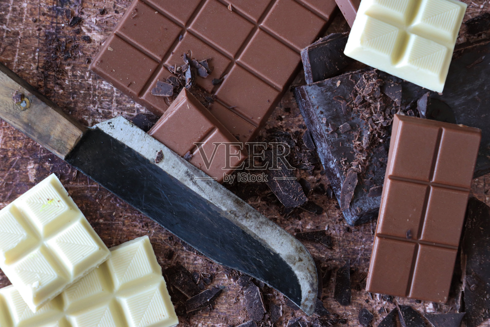 全帧图像的厨房刀与巧克力棒，牛奶巧克力块，白巧克力方块和黑巧克力块磨碎的棕色背景，抬高视图照片摄影图片