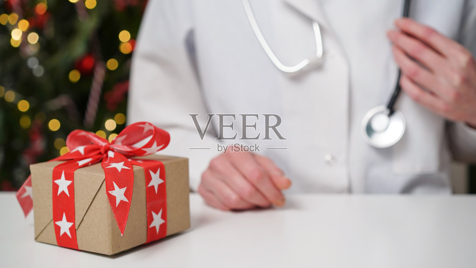 圣诞节和新年医疗横幅概念。穿着白大褂的女医生坐在她的办公桌前，桌子上有一个红色蝴蝶结的礼盒，背景是装饰精美的圣诞树。照片摄影图片