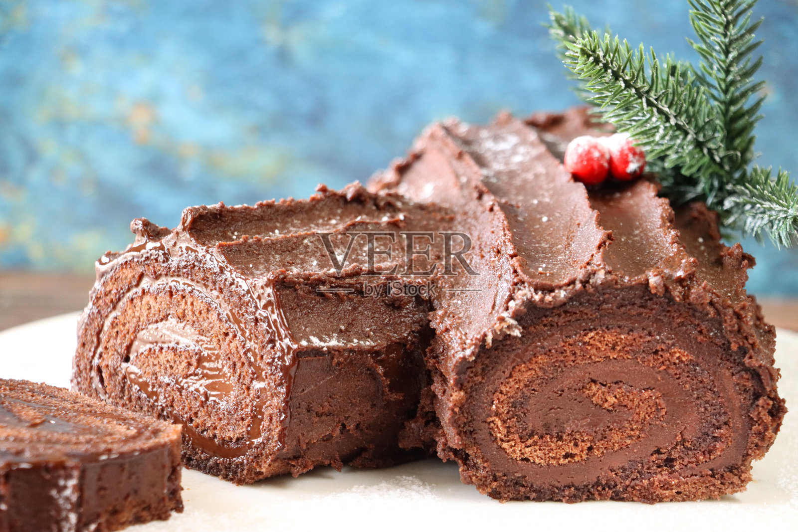 白色盘子上装饰的巧克力圣诞原木蛋糕的特写图像，瑞士卷海绵，巧克力甘纳许填充，天然树皮图案蚀刻成巧克力奶油糖霜，云杉针和红色浆果，蓝色背景，重点在前景照片摄影图片