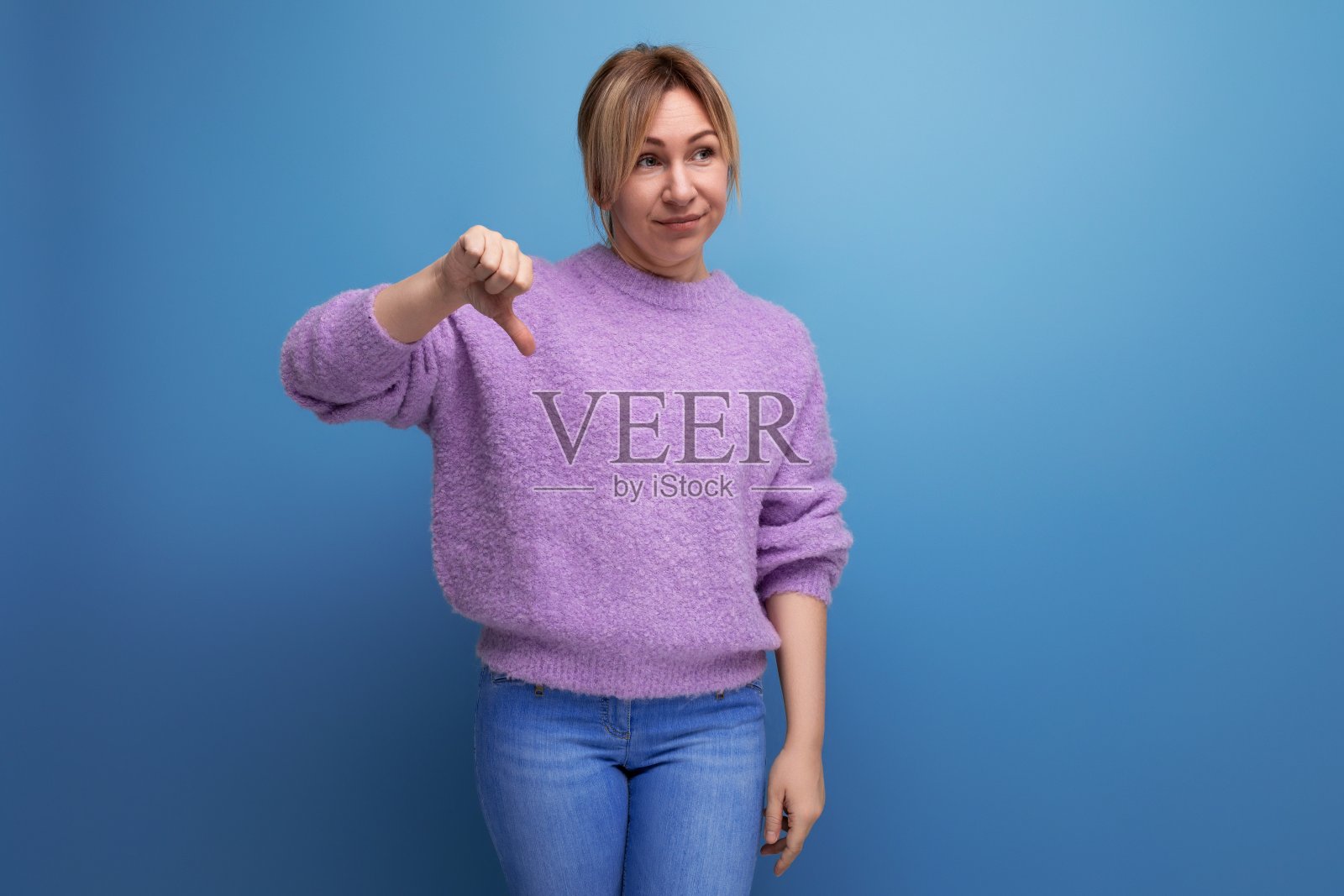 迷人的令人愉快的金发年轻女子的肖像在紫色毛衣与不喜欢的手势在蓝色背景与复制空间照片摄影图片