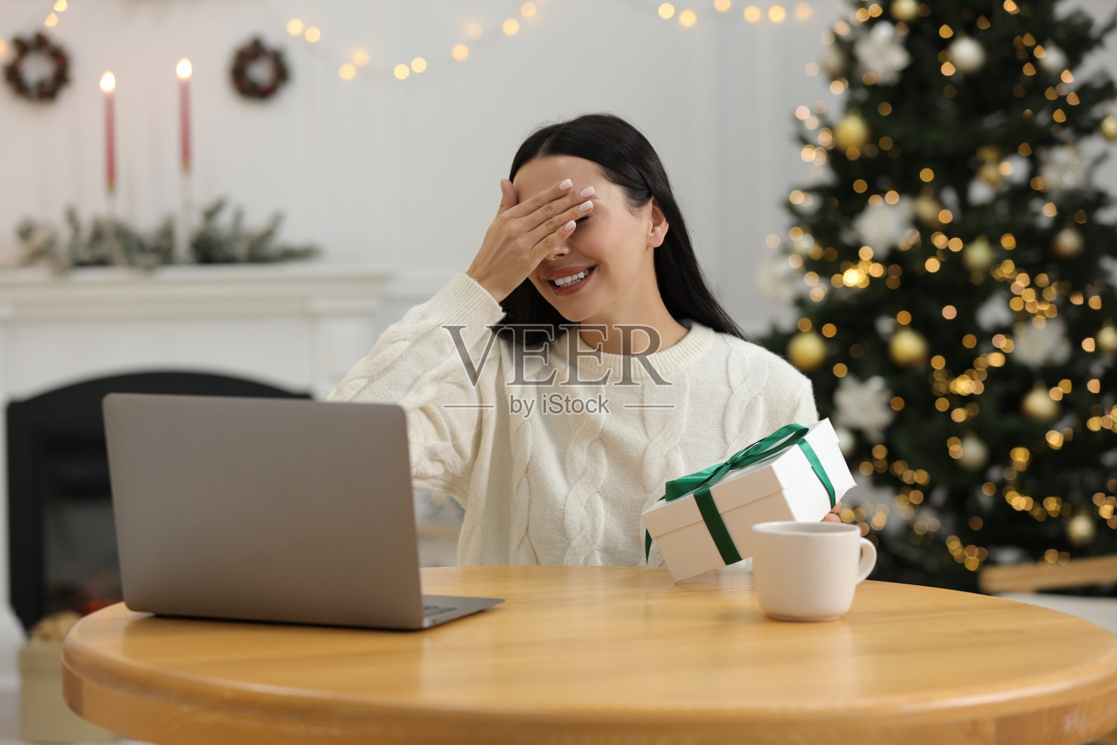 通过邮件交换礼物，在网上庆祝圣诞节。在家里视频通话时，微笑的女人在打开礼盒前遮住眼睛照片摄影图片