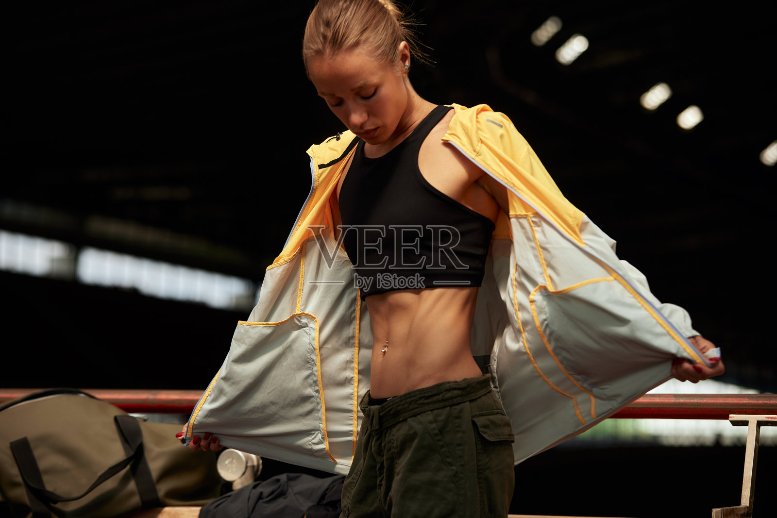 年轻的健身女孩正准备在体育馆锻炼。迷人苗条的金发女郎穿着黄灰色运动夹克、黑色上衣和卡其色工装裤换了衣服。积极的生活方式。照片摄影图片