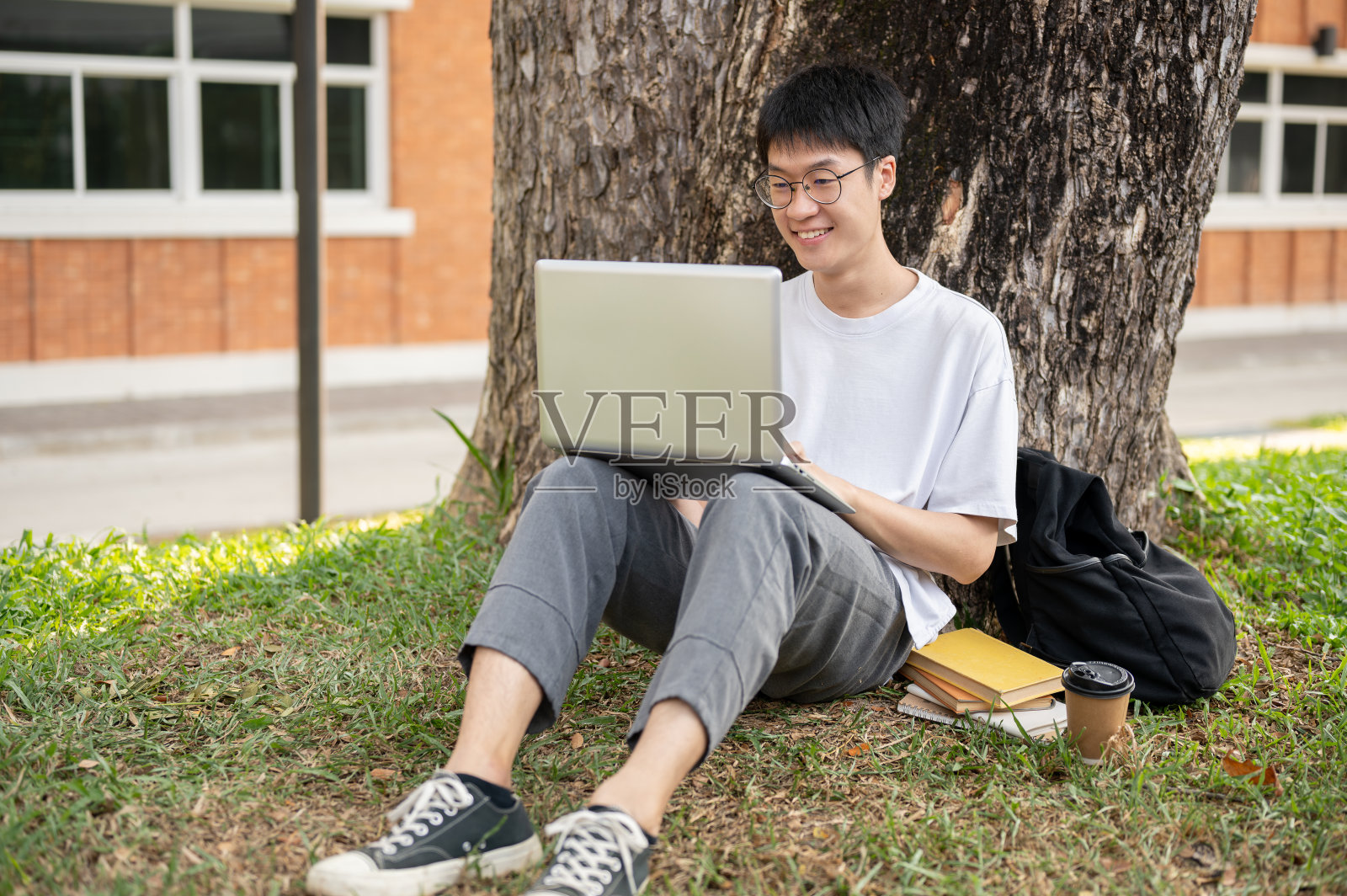 一位年轻快乐的亚洲男大学生正在校园公园的树下使用他的笔记本电脑。照片摄影图片