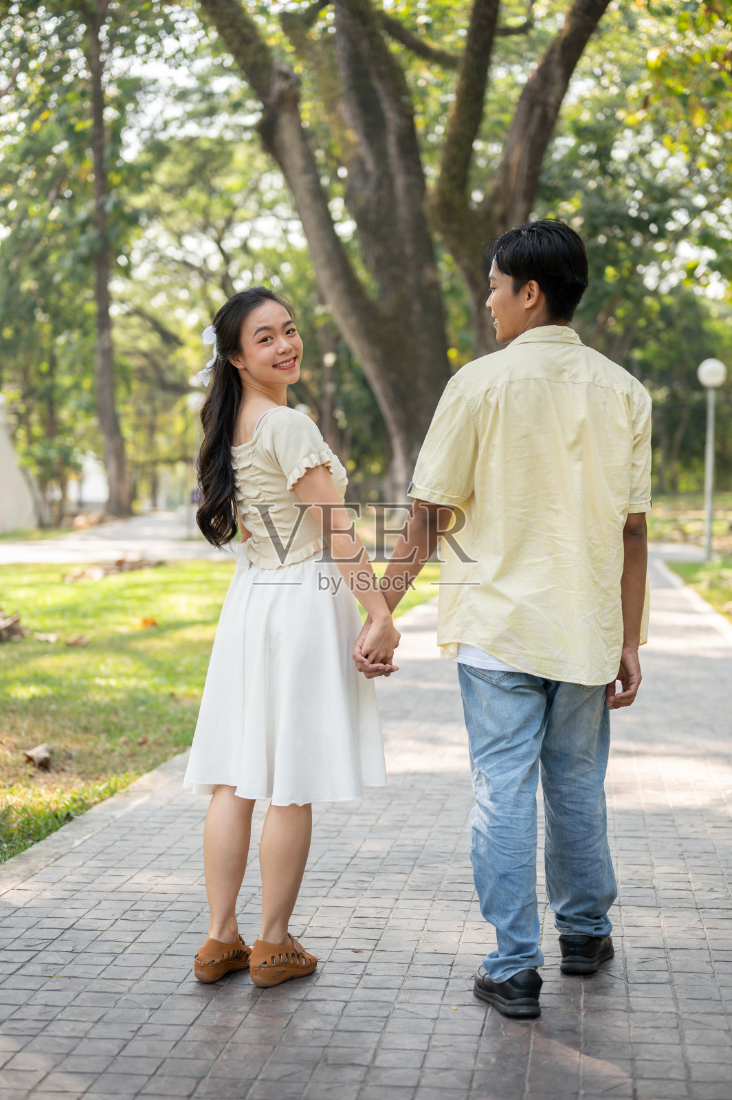 一位年轻漂亮的亚洲女性牵着男友的手在绿色的公园里散步。照片摄影图片