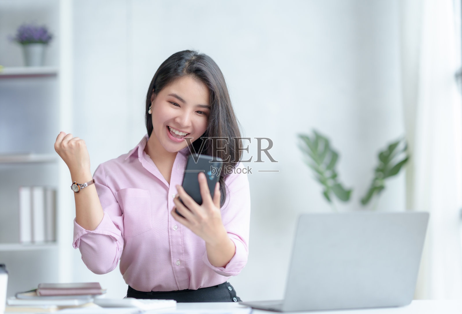 快乐的年轻亚洲女商人成功兴奋举起手，用智能手机庆祝。照片摄影图片