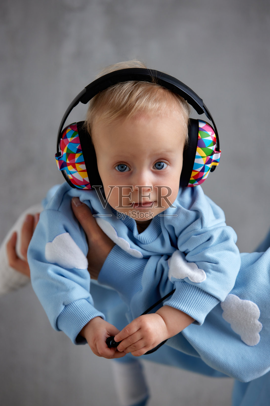 一位快乐的年轻母亲抱着她的宝贝孩子。孩子用灰色的眼睛看着镜头，头上戴着欢快的彩色DJ耳机。照片摄影图片