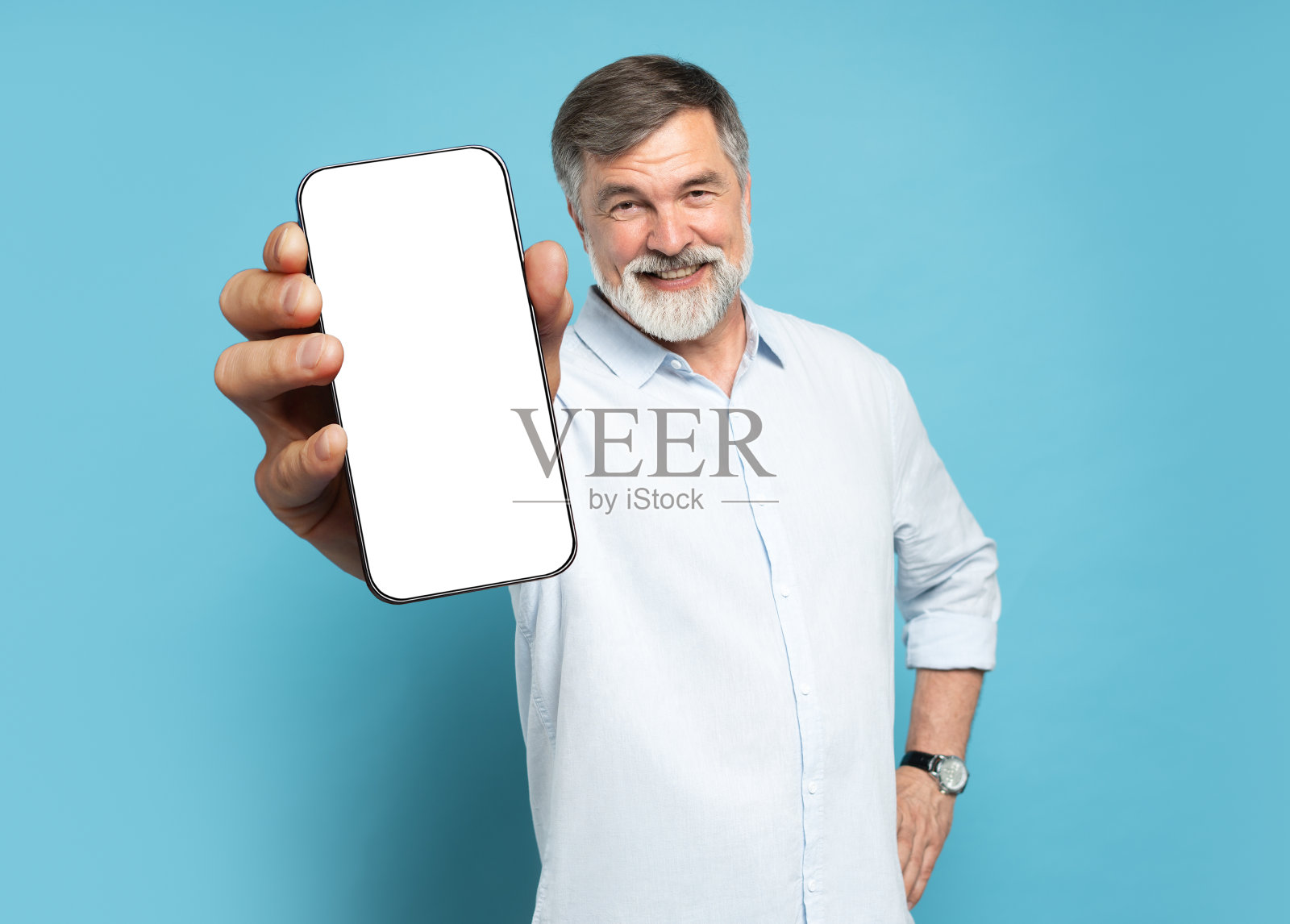 英俊兴奋的成熟男子指着空的智能手机屏幕，在浅蓝色的工作室背景下摆姿势，对着镜头微笑。看看这个，手机显示屏模型照片摄影图片