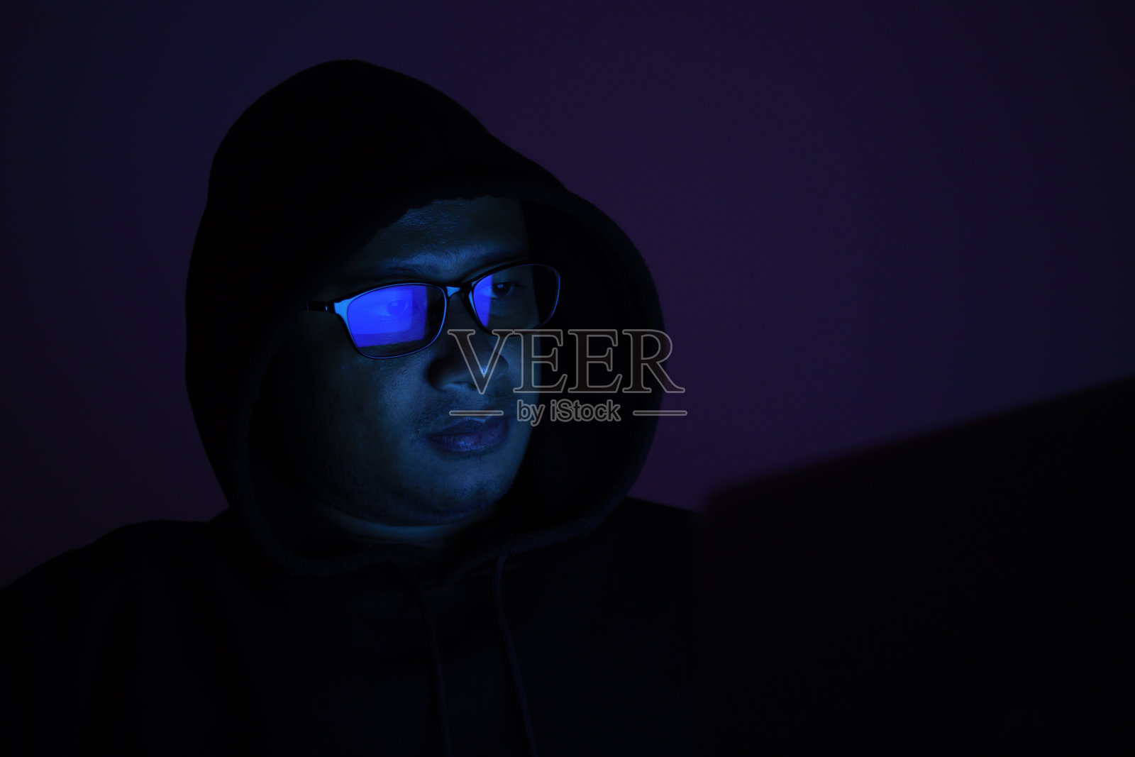 一名戴着眼镜、戴着连帽衫、在黑暗中蒙住头的男性黑客正在侵入受害者的金融系统。数据盗窃，网络欺诈，暗网和网络安全概念。照片摄影图片