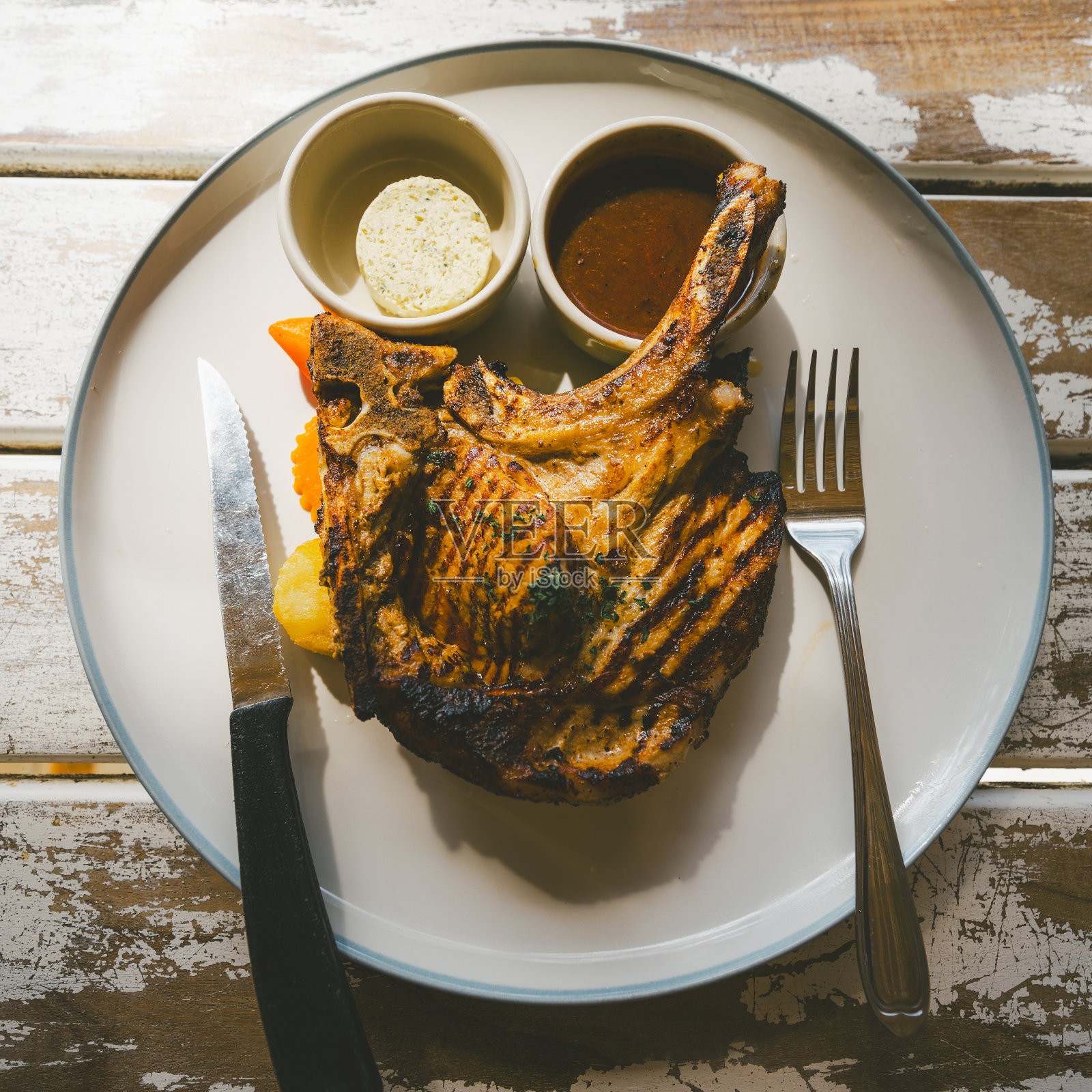 白色盘子里的烤猪排和蔬菜，白色木桌上有一碗酱汁和奶酪球。照片摄影图片