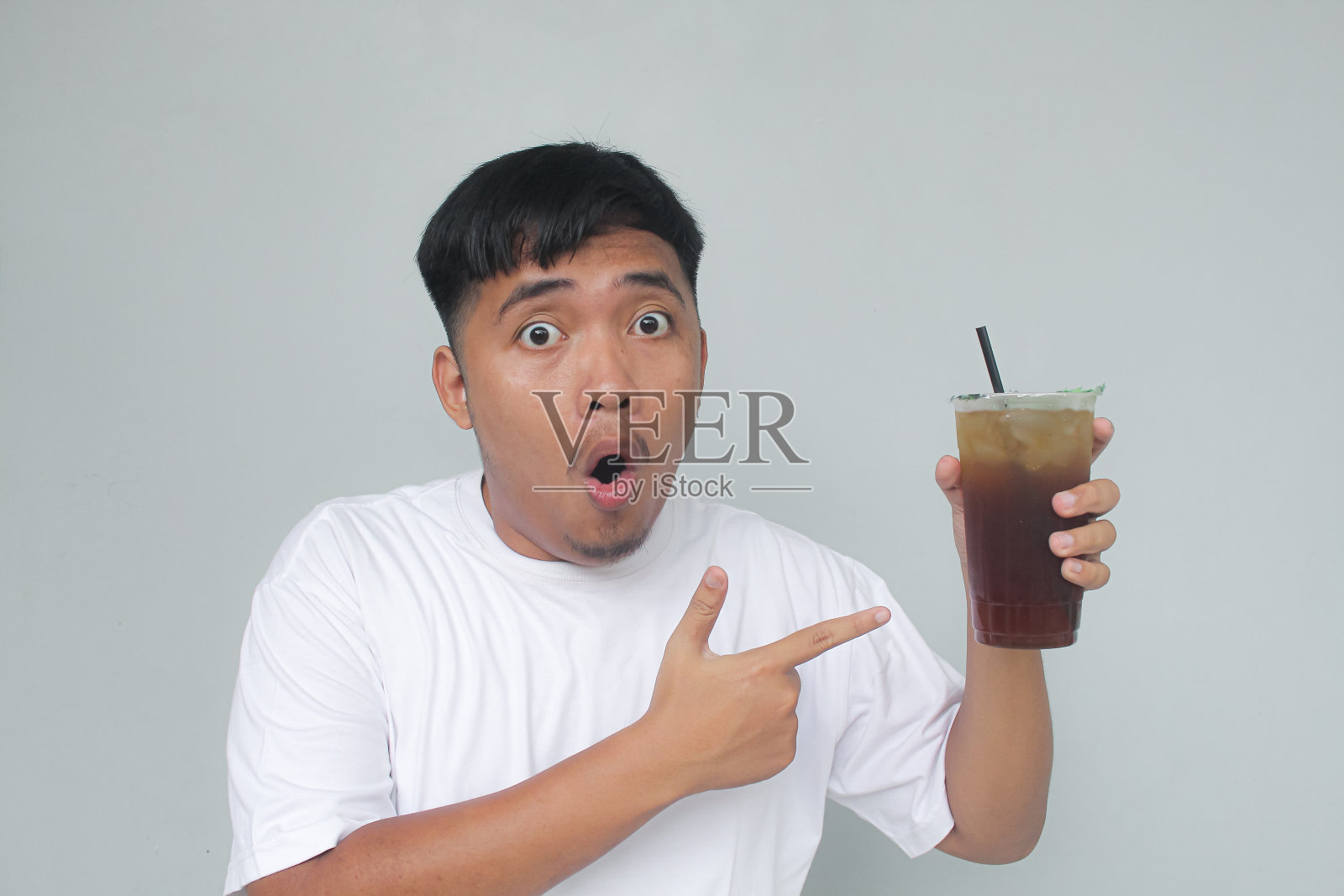一个亚洲年轻人拿着冰茶，用手指着，脸上带着“哇”的表情。照片摄影图片