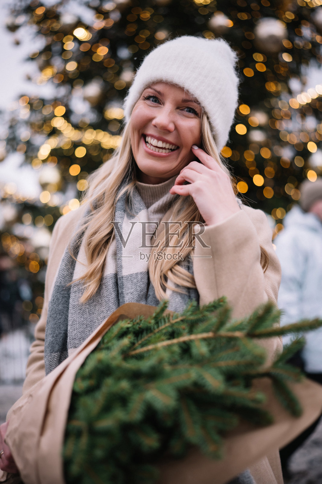身穿米色大衣的年轻金发女子手持一束新鲜的冷杉树枝走在城市里照片摄影图片