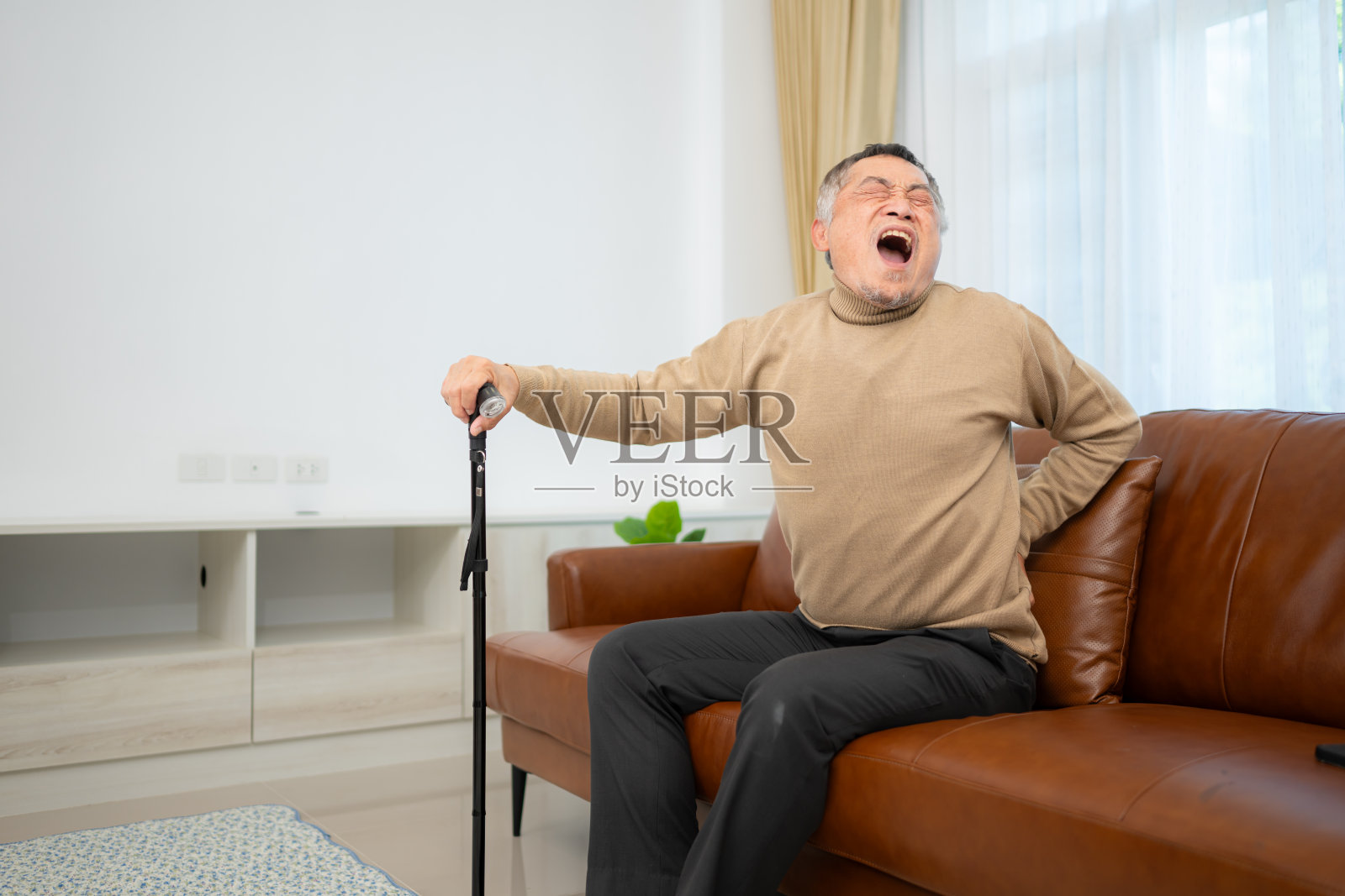 由于背痛，一位老人拄着拐杖坐在家里客厅的沙发上。照片摄影图片