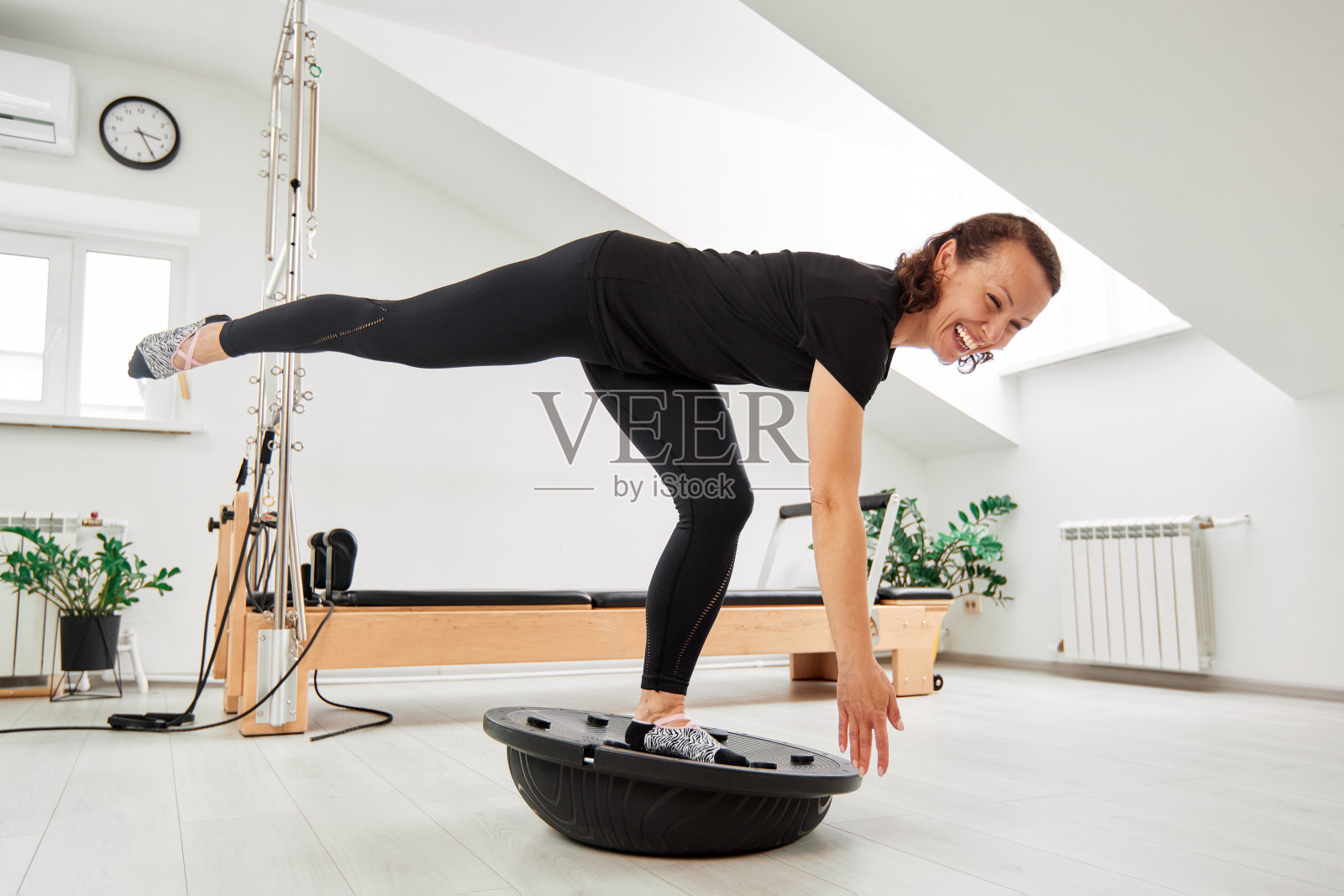 一个女人在一间明亮的工作室里和改革家们一起做普拉提。一位身材修长、柔韧的黑发女子身穿黑色紧身衣，在大脑半球进行锻炼，以平衡和增强肌肉。健康的生活方式。照片摄影图片