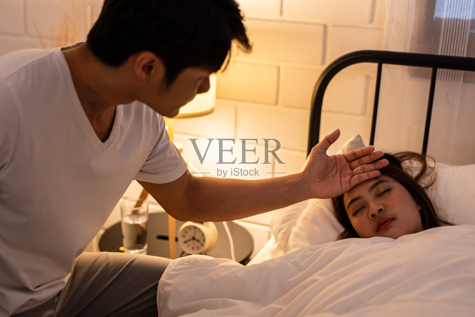 亚洲的丈夫在晚上照顾躺在床上生病的妻子。有吸引力的年轻人支持和帮助美丽的困倦的女朋友感到发烧和咳嗽在家里的卧室。照片摄影图片