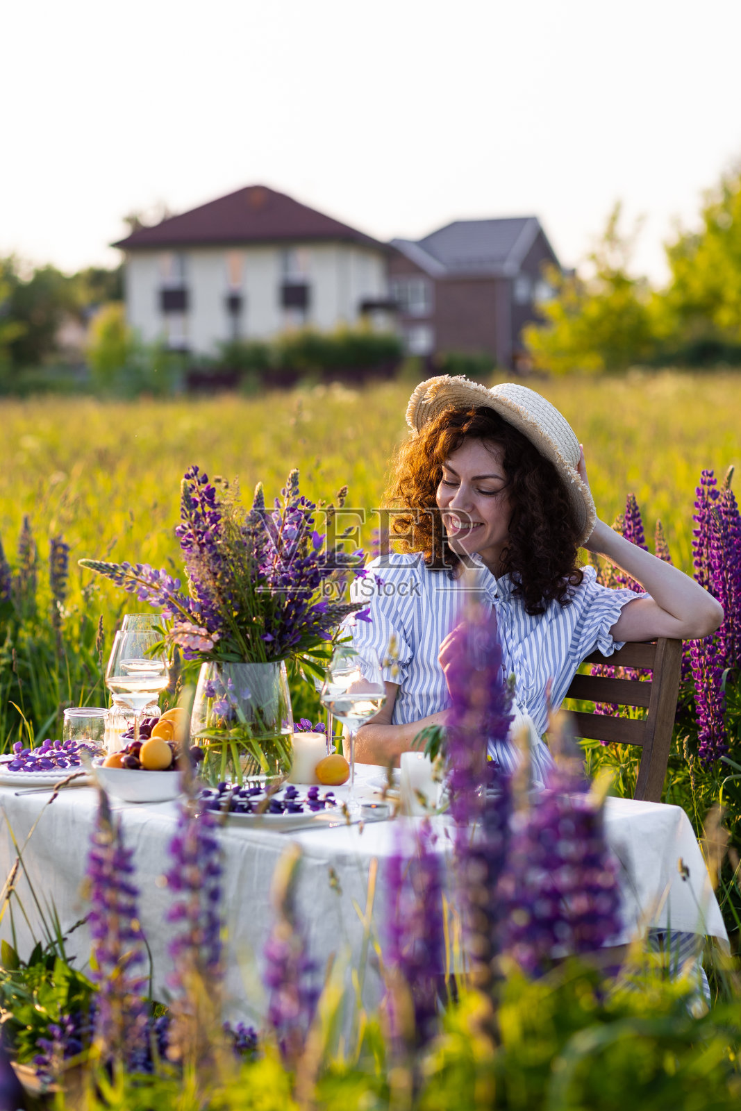 黄金时刻，日落，夏天。美丽快乐的年轻女子在草地上为户外活动安排桌子，采集野花。婚礼或浪漫的约会装饰在外地与紫色羽扇豆照片摄影图片