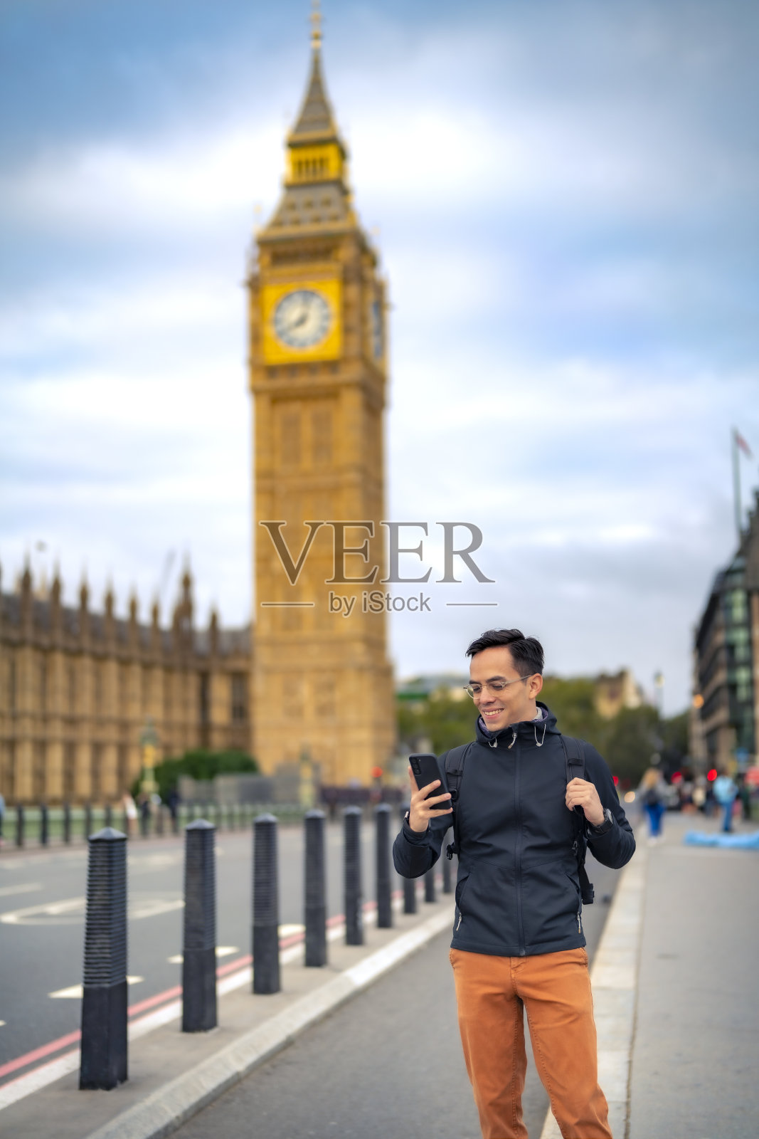 身着休闲装、戴着眼镜的年轻拉丁男游客，微笑着站在大本钟附近的桥上，对着钟楼看手机照片摄影图片