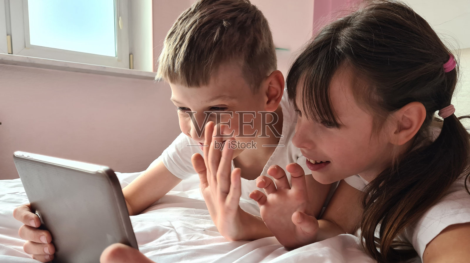 微笑的小孩子，男孩和女孩躺在床上看着屏幕的平板电脑。照片摄影图片