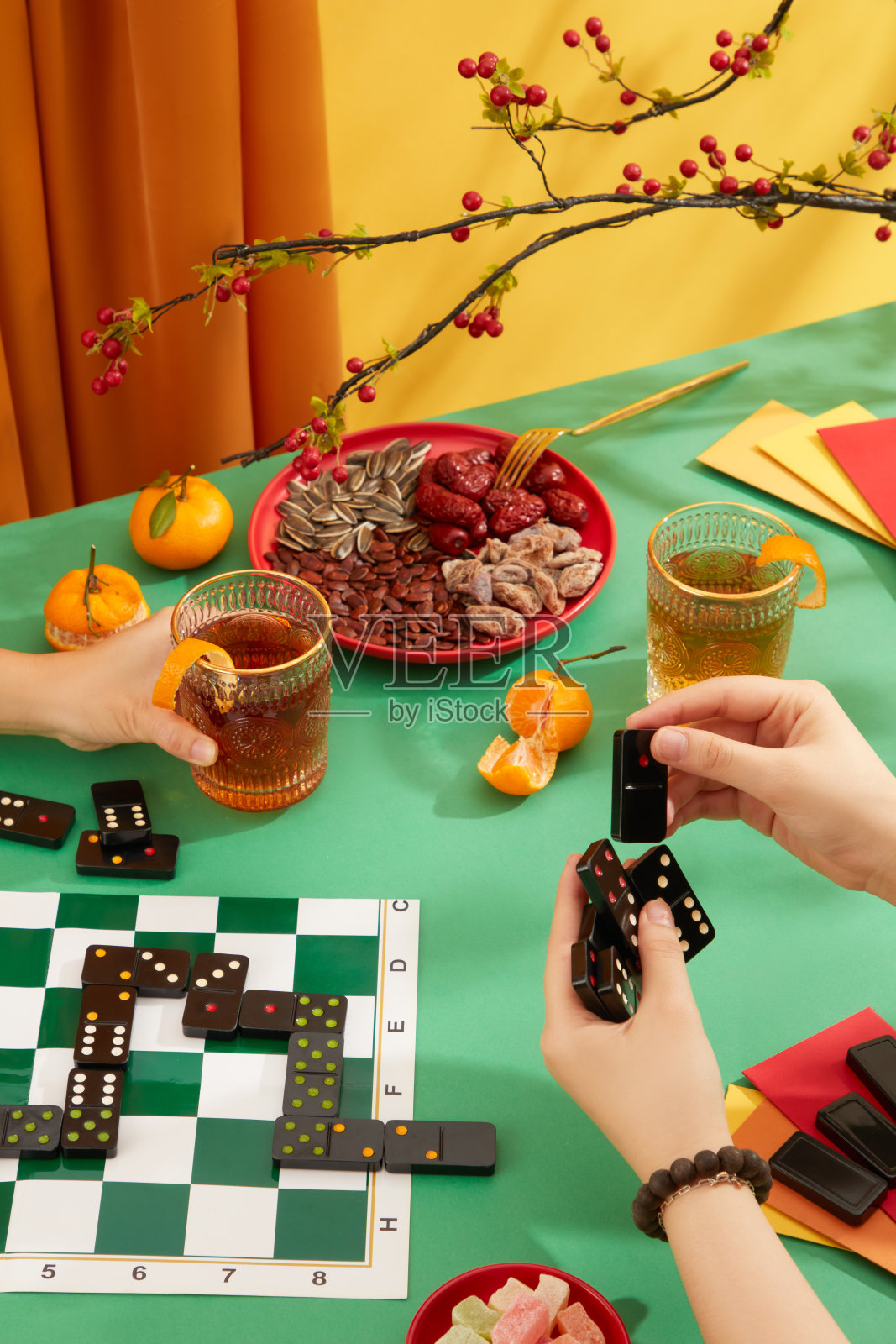 两个人在桌子上玩多米诺骨牌，周围是一盘干果和果酱、橘子、两杯碳酸水和红包。春节的气氛是热闹的。照片摄影图片