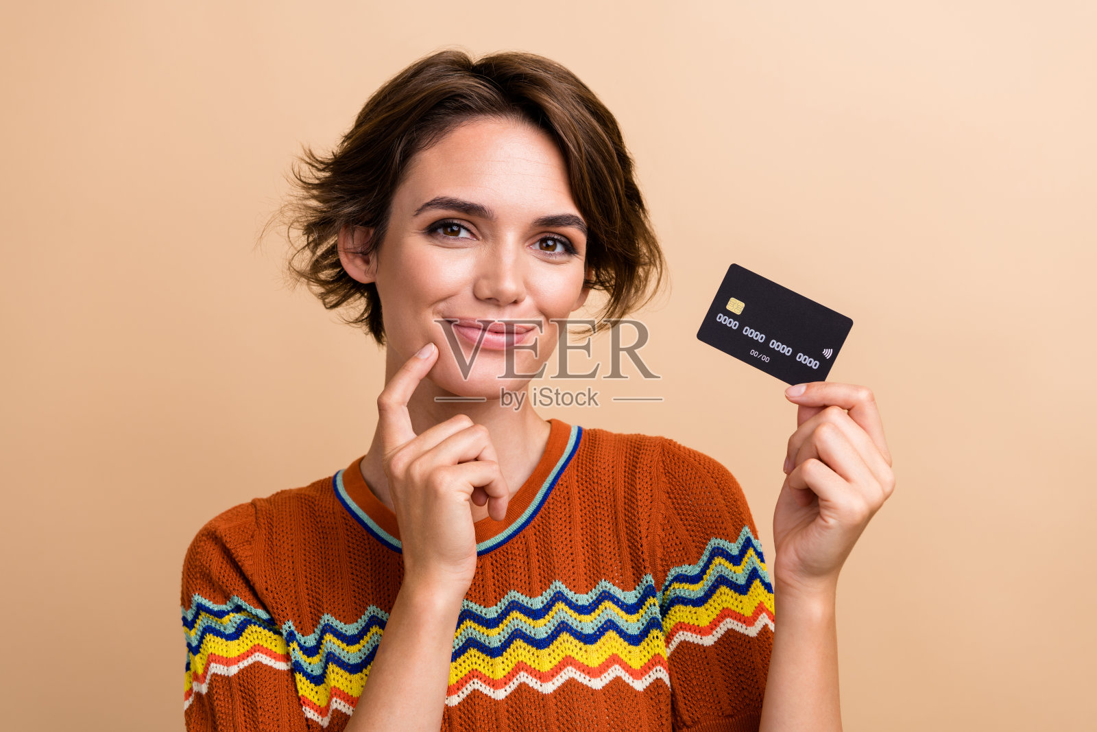 照片可爱的积极向上的女孩手指触摸下巴，手臂持有借记卡孤立的米色背景照片摄影图片