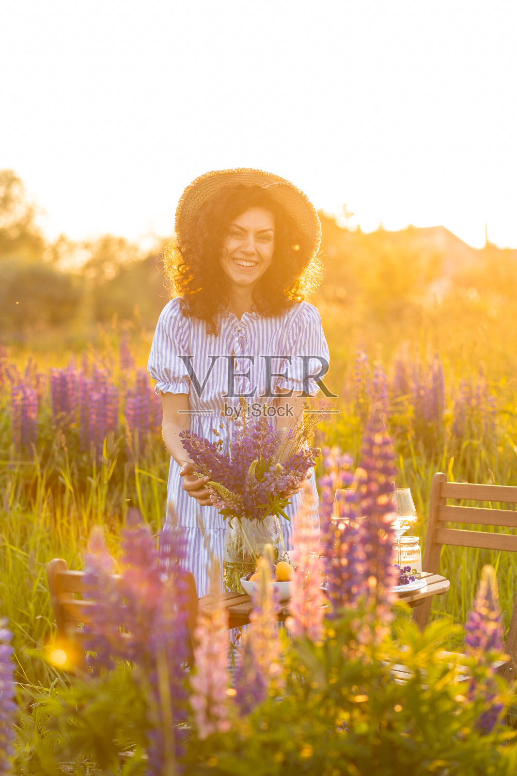 黄金时刻，日落，夏天。美丽快乐的年轻女子在草地上为户外活动安排桌子，采集野花。婚礼或浪漫的约会装饰在外地与紫色羽扇豆照片摄影图片