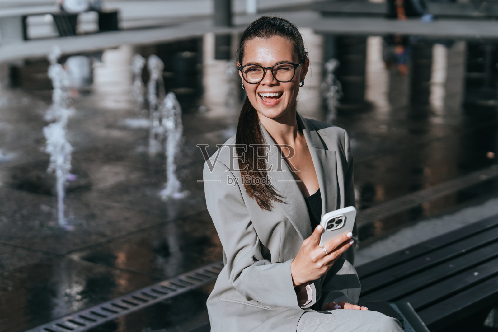 笑着的戴眼镜的职业女性坐在长凳上，用她的智能手机，背景是饮水机照片摄影图片