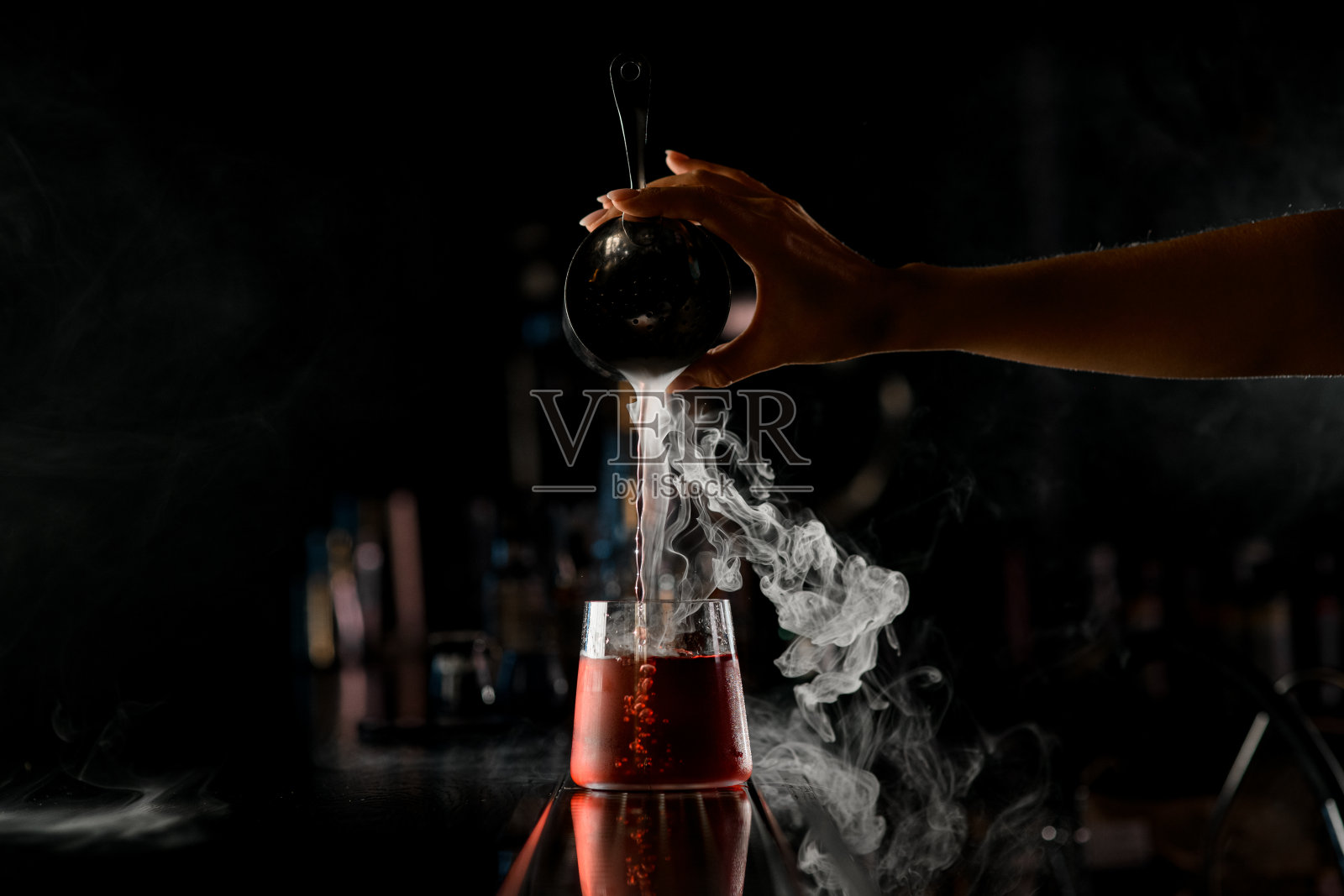 女调酒师的手准确地把热气腾腾的液体从钢摇壶里倒进吧台前的酒杯里照片摄影图片