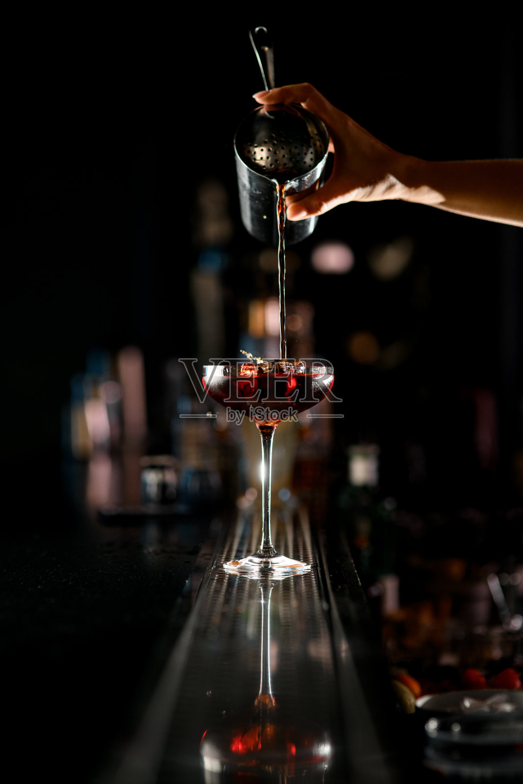 垂直拍摄的女调酒师的手将红色鸡尾酒倒入杯中，吧台上放着冰块照片摄影图片