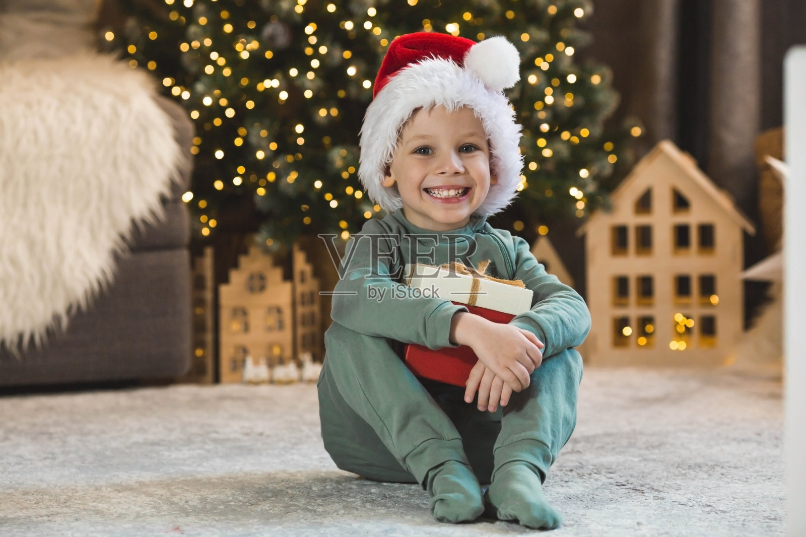 一个漂亮的小男孩坐在圣诞树下，手里拿着一个红盒子。一份新年礼物。庆祝2024年圣诞节和新年的概念。照片摄影图片